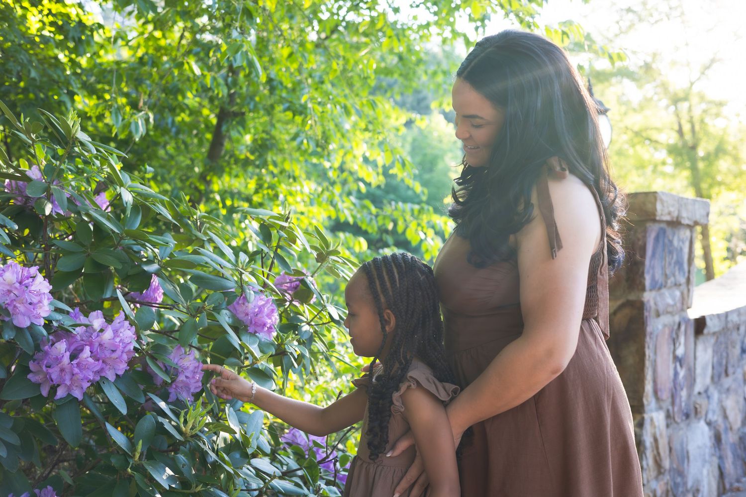 A mother and daughter admire purple rhododendron flowers in a lush garden setting.