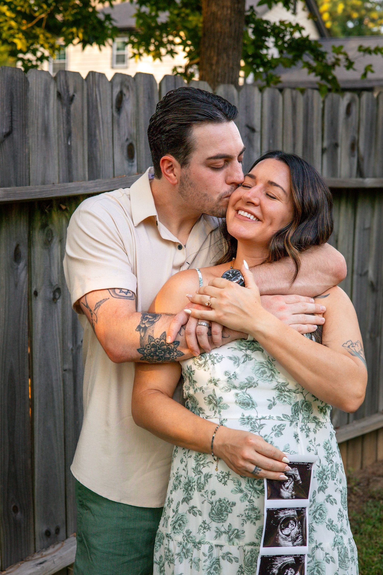Romantic couple embraces in front of wooden fence while holding ultrasound photos in backyard setting.