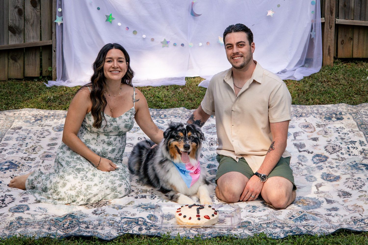 A couple sits on a white blanket backdrop with decorative lights and confetti in an outdoor setting.