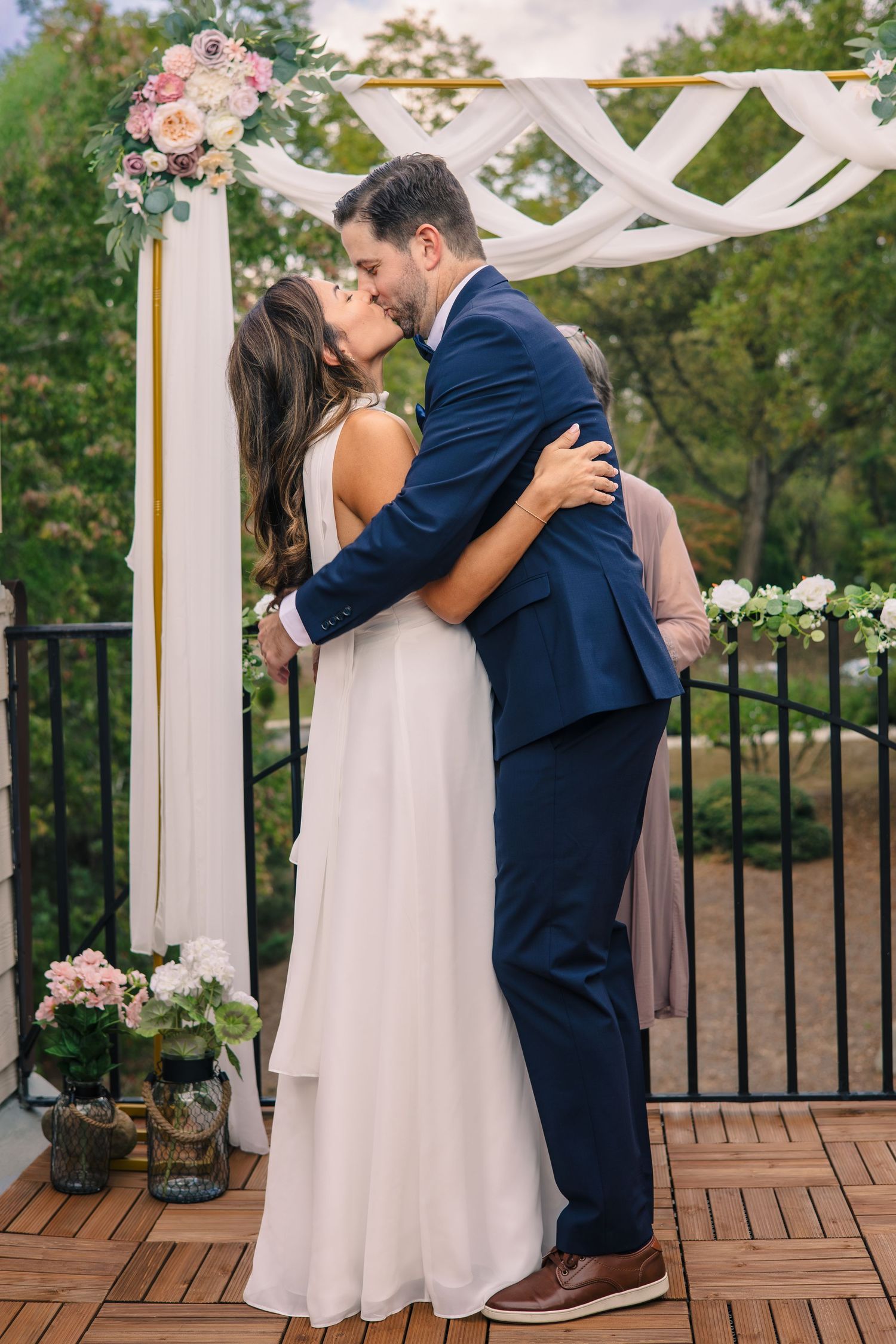 Couple shares a romantic first kiss under a white pergola decorated with pink flowers during outdoor wedding ceremony.
