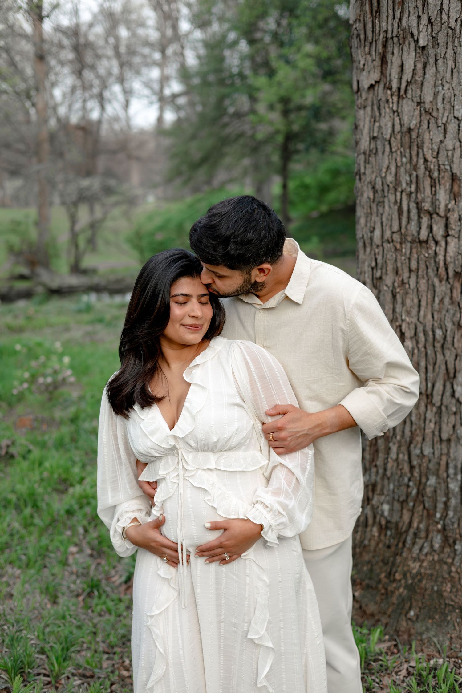 A pregnant woman in a white dress is embraced by her partner in a natural outdoor setting.