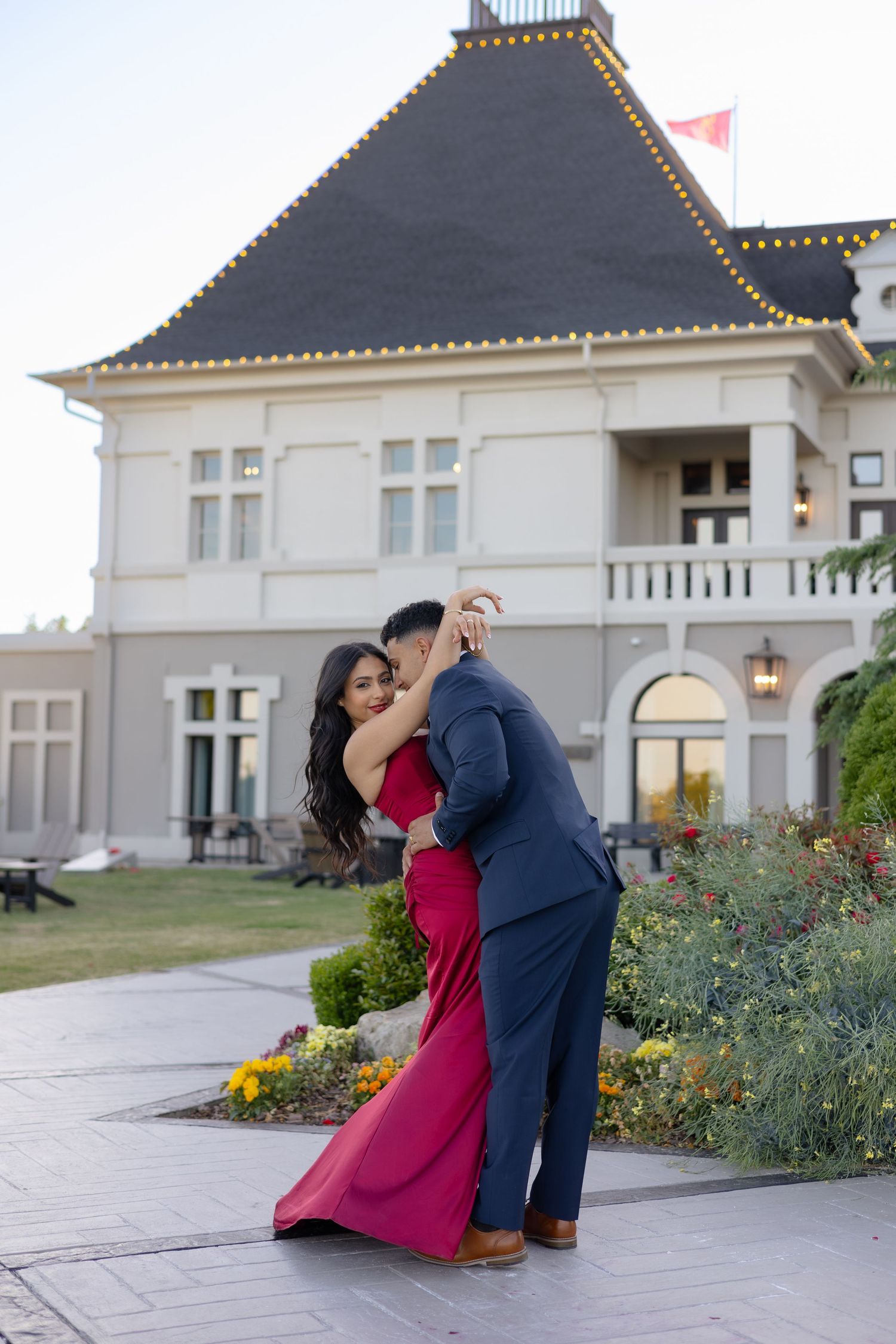 Couple dancing romantically outside elegant mansion at dusk, woman in red gown and man in navy suit.