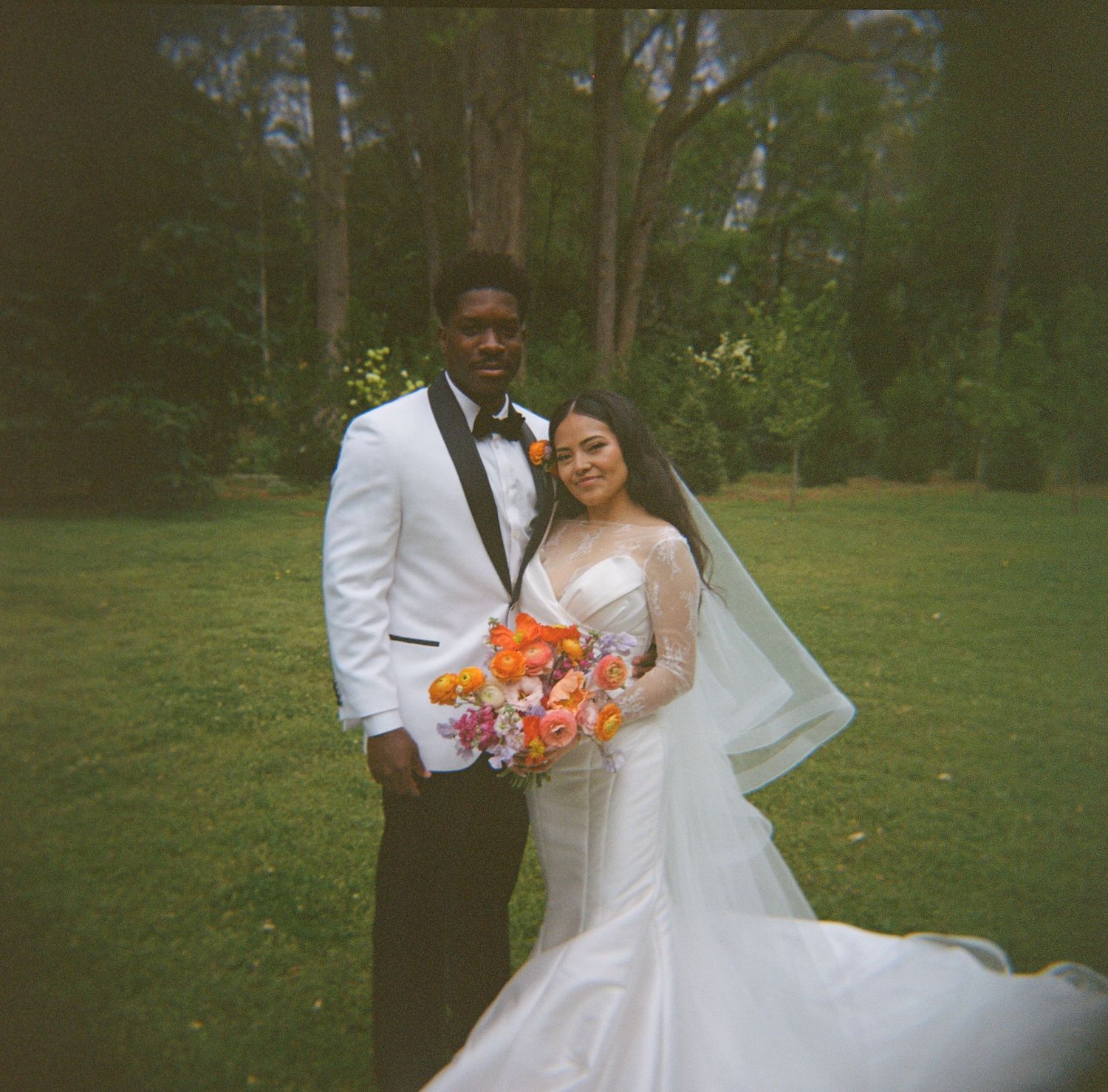 Bride and groom pose outdoors on lush green lawn, she holds vibrant orange bouquet, wearing white gown with veil.