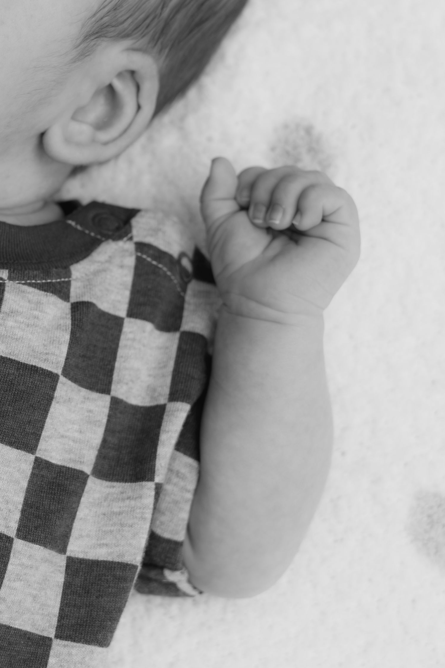 Black and white photo of a baby in a checkered onesie, tiny fist raised, lying on a soft white surface.