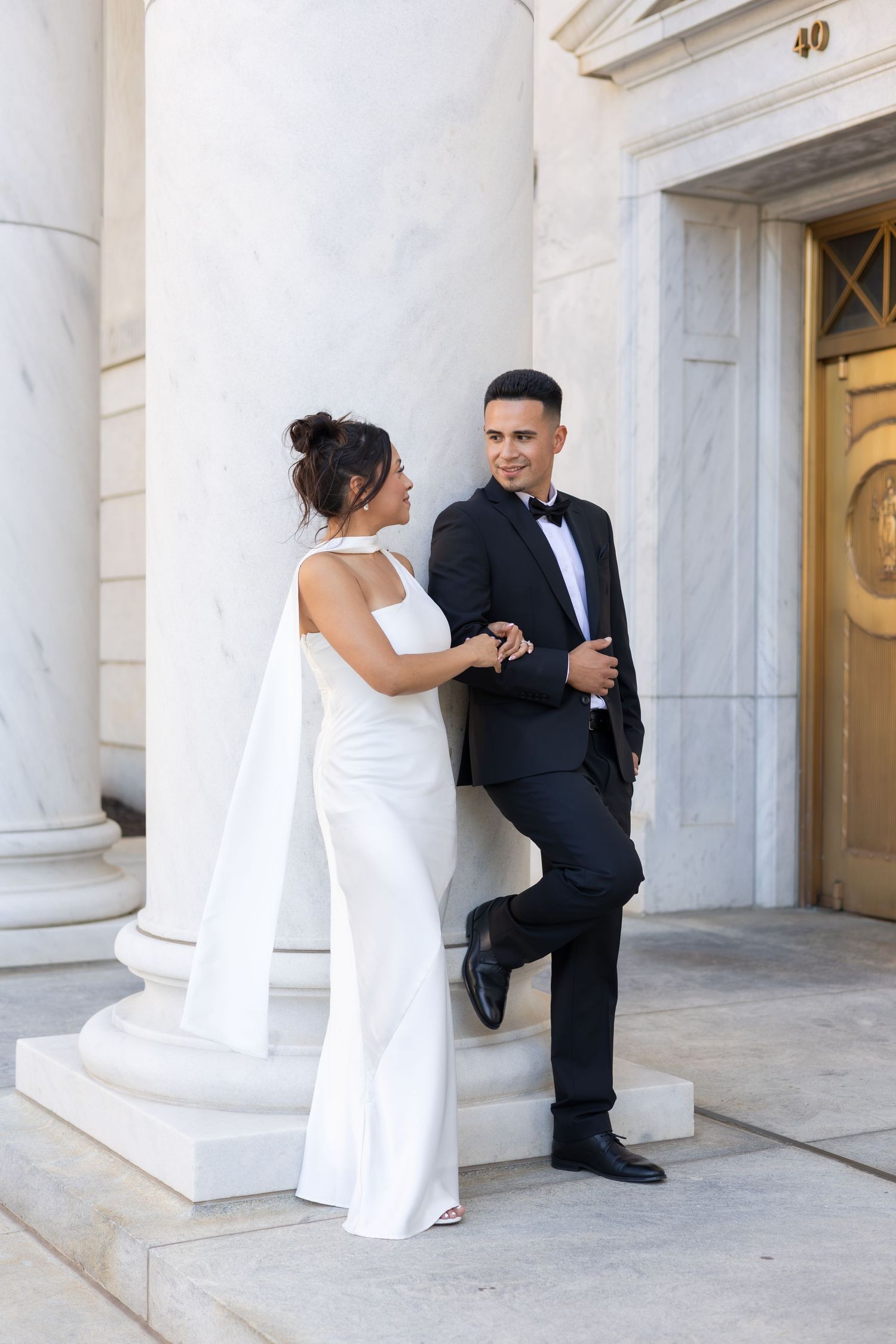 Elegant couple in wedding attire posing by marble columns, bride in white cape gown, groom in black tuxedo.