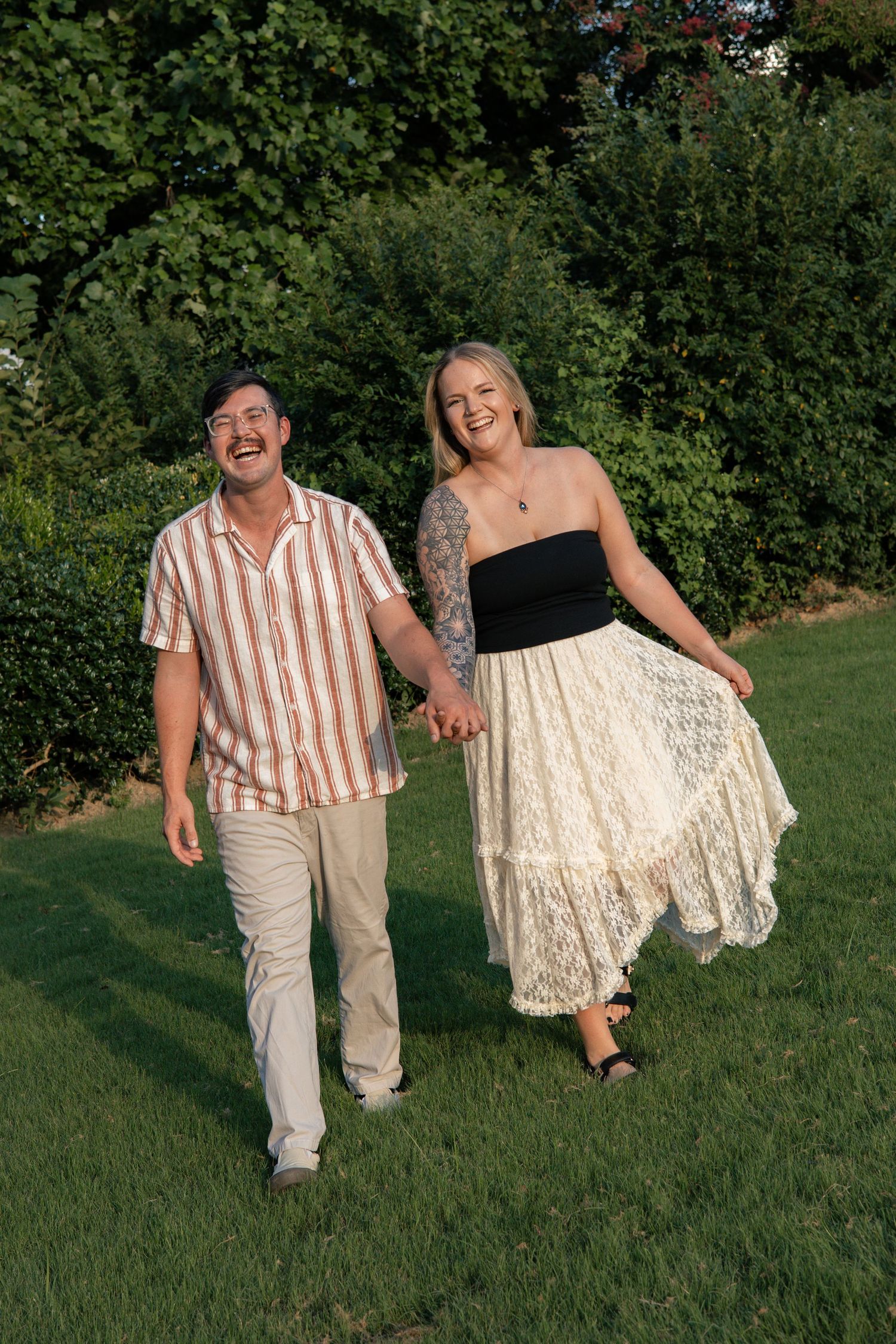 A couple laughs together while walking hand in hand through a garden setting.