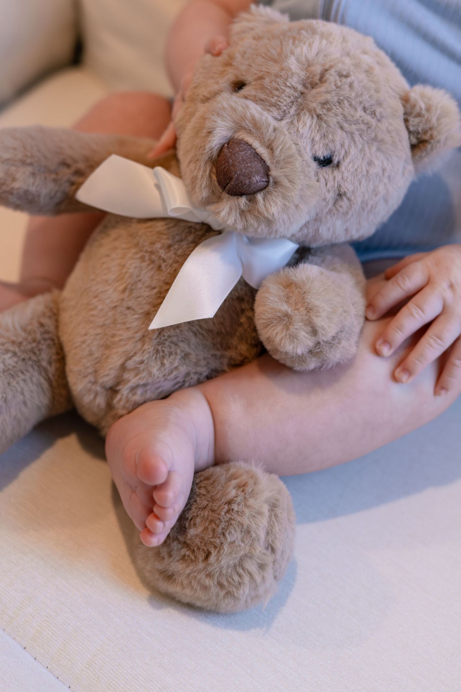 Soft plush teddy bear with white bow resting on light colored fabric.