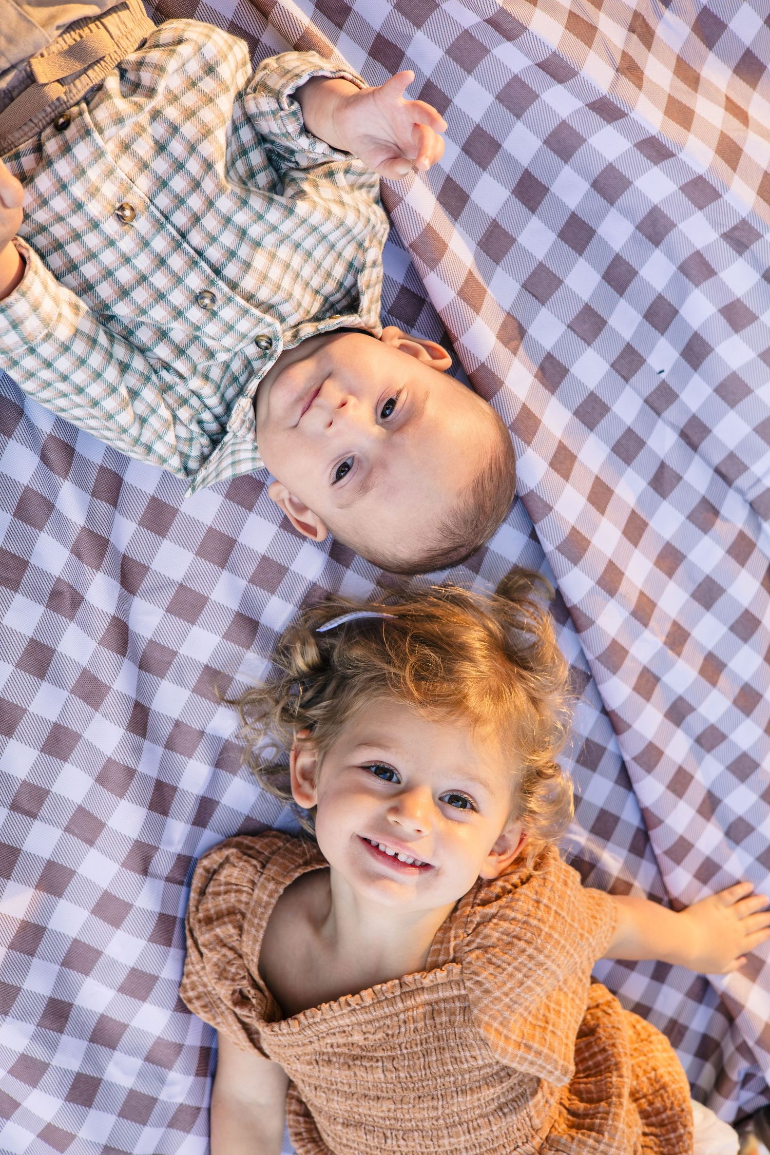 Two children lying on a checkered picnic blanket smiling at the camera.