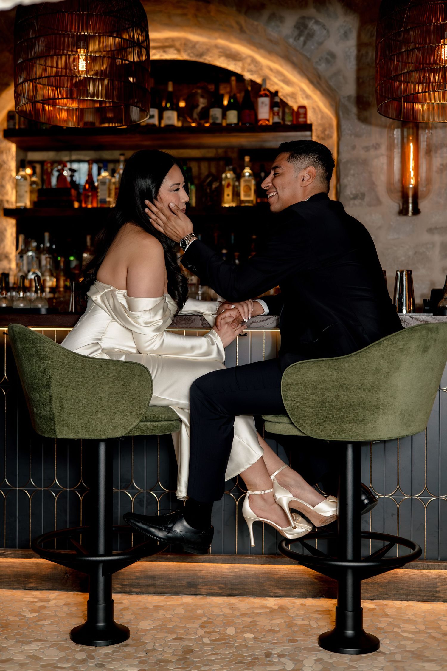 Couple sharing a romantic moment at an upscale bar, woman in white satin dress and heels, man in black suit.