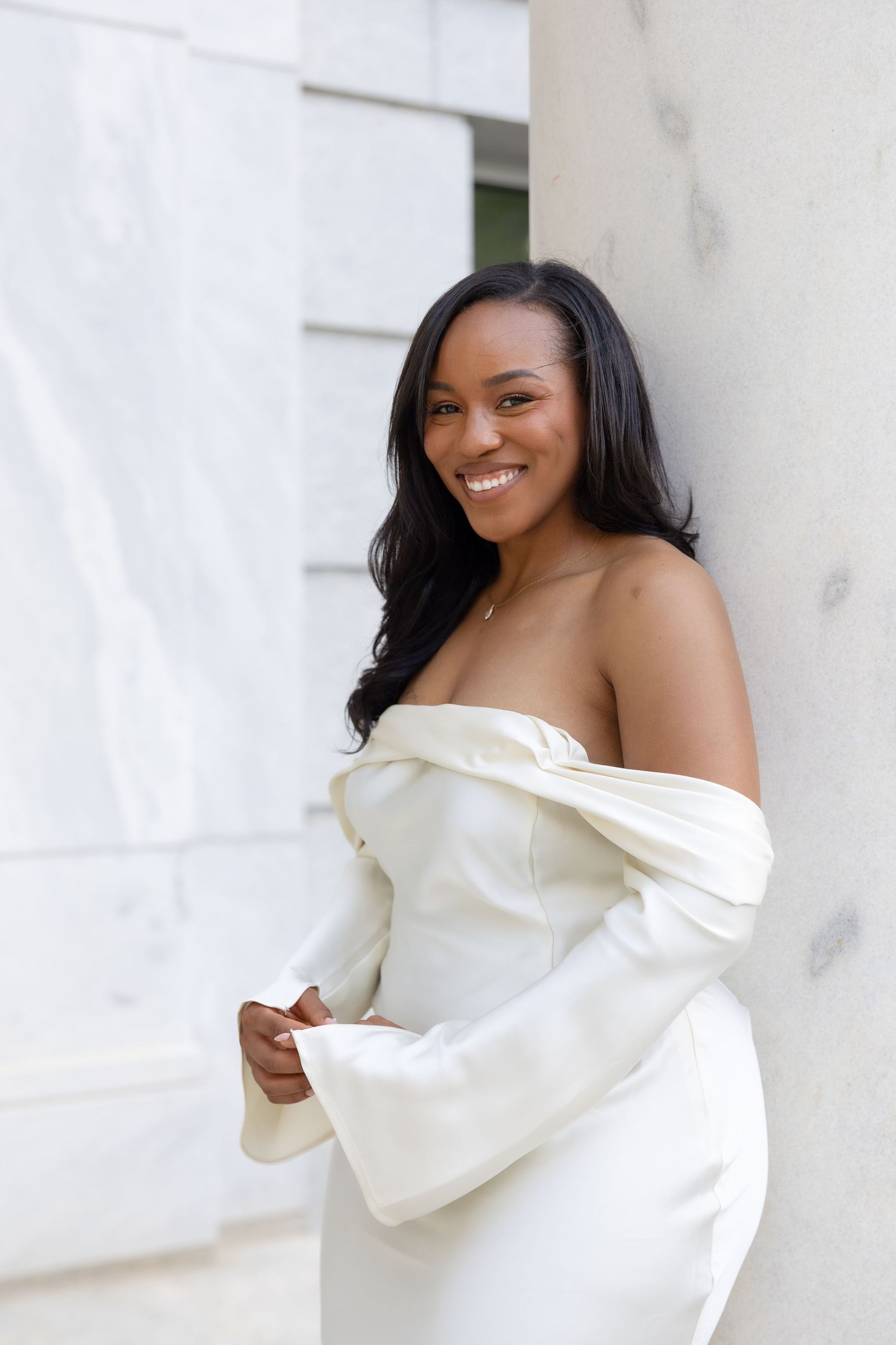 Smiling Black woman in elegant off-shoulder white gown with bell sleeves, leaning against marble column outdoors.