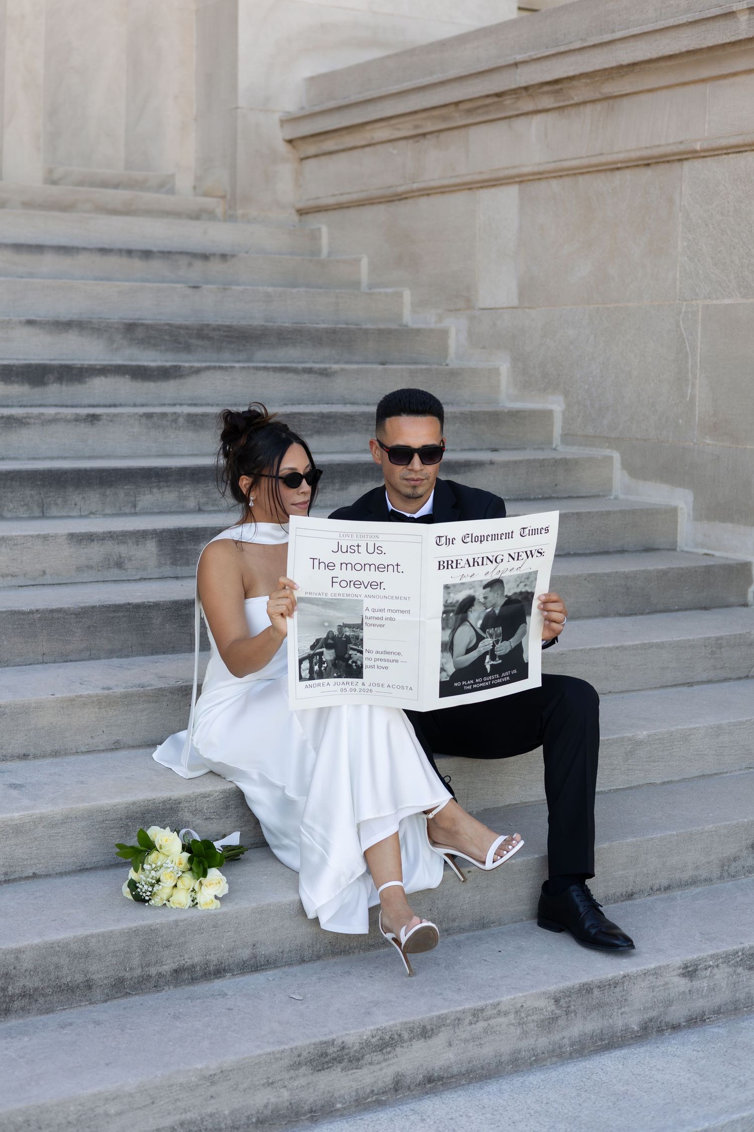 Newlywed couple in wedding attire sitting on stone steps reading a newspaper together, bride holding white bouquet.