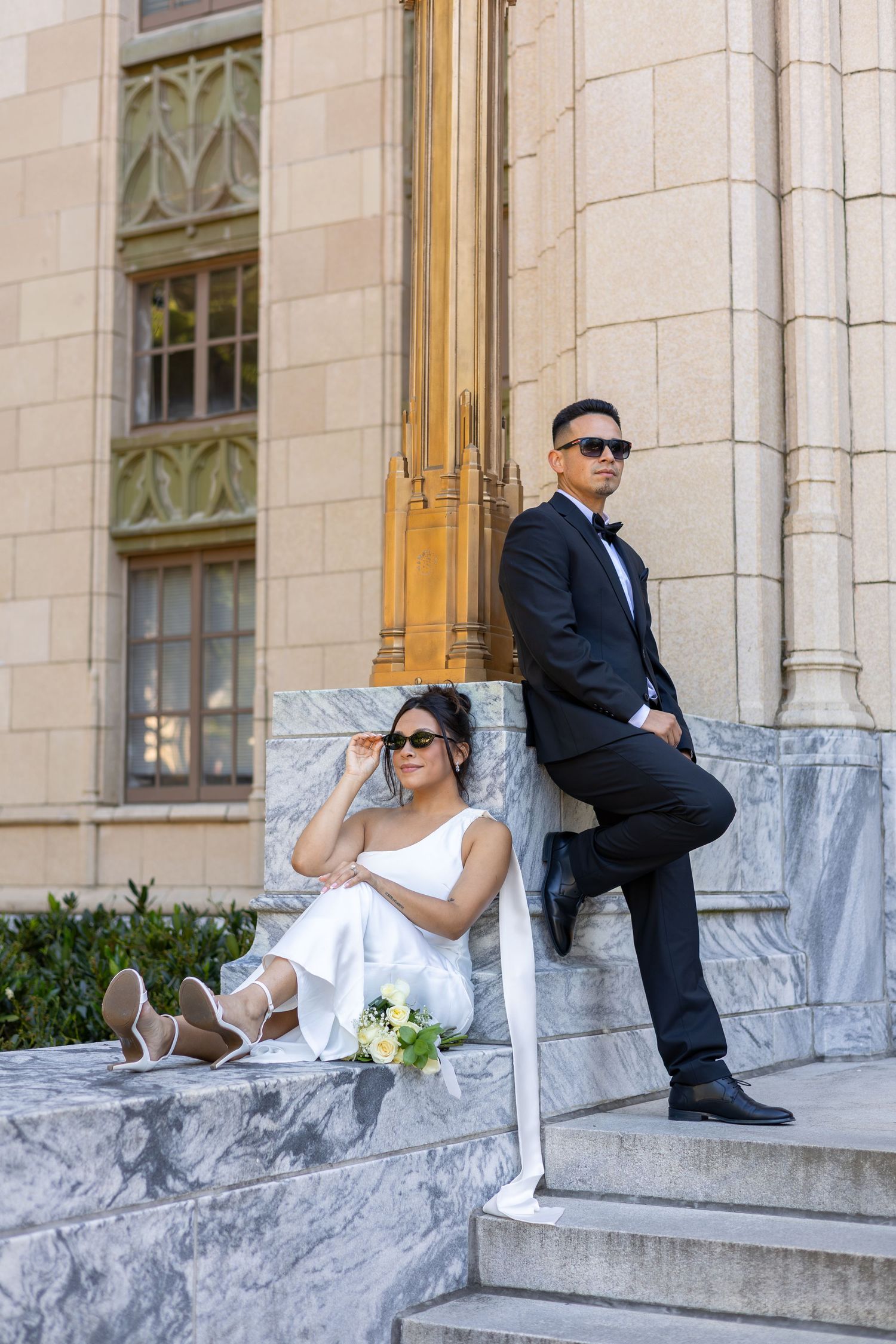 Bride and groom posing on marble steps of elegant building, she sits holding bouquet, he stands leaning on golden column.