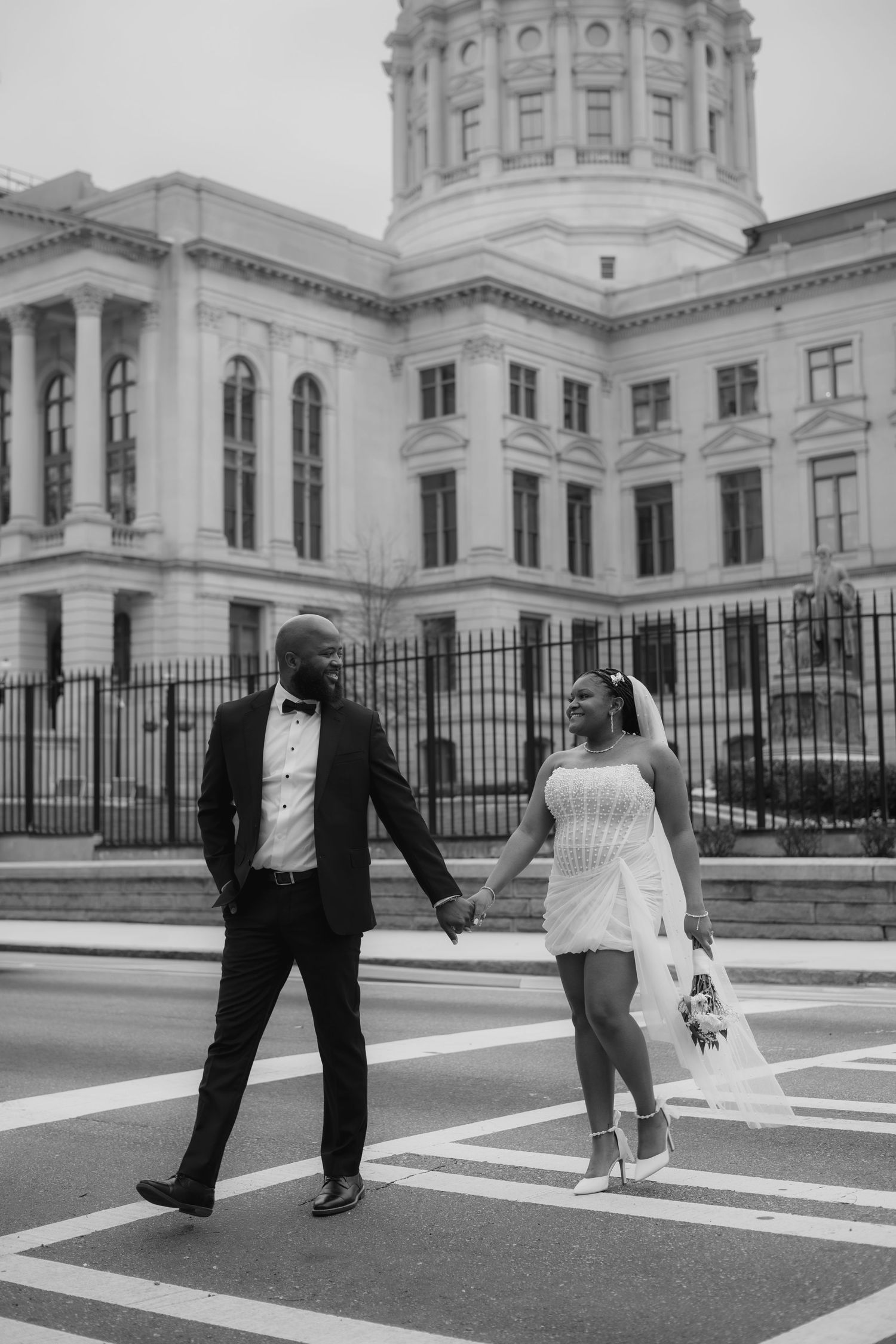 A couple in wedding attire playfully poses in front of a grand government building with white architecture and dome.