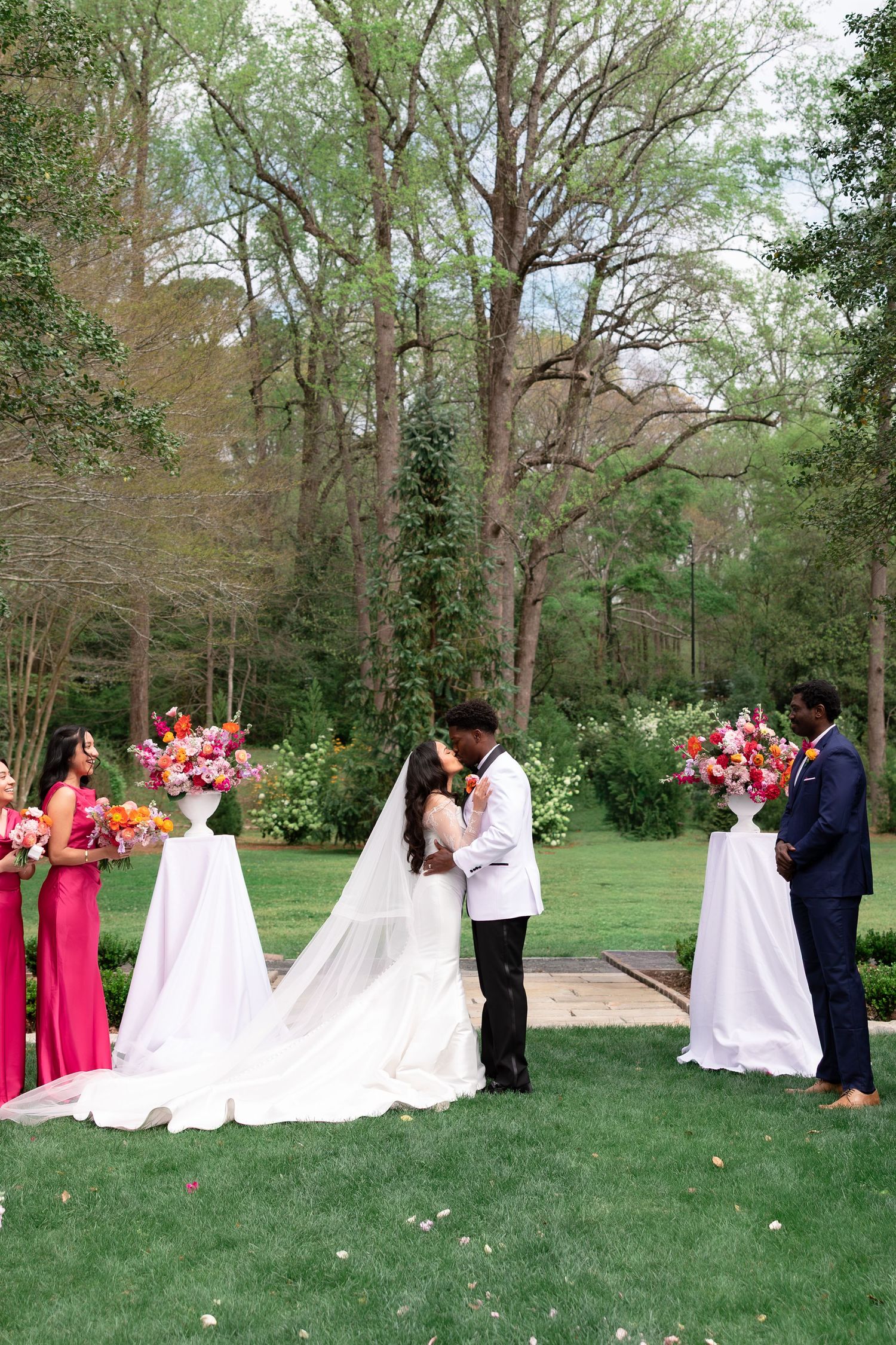 Bride and groom share first kiss at outdoor garden wedding ceremony with pink floral arrangements and wedding party on lush green lawn.