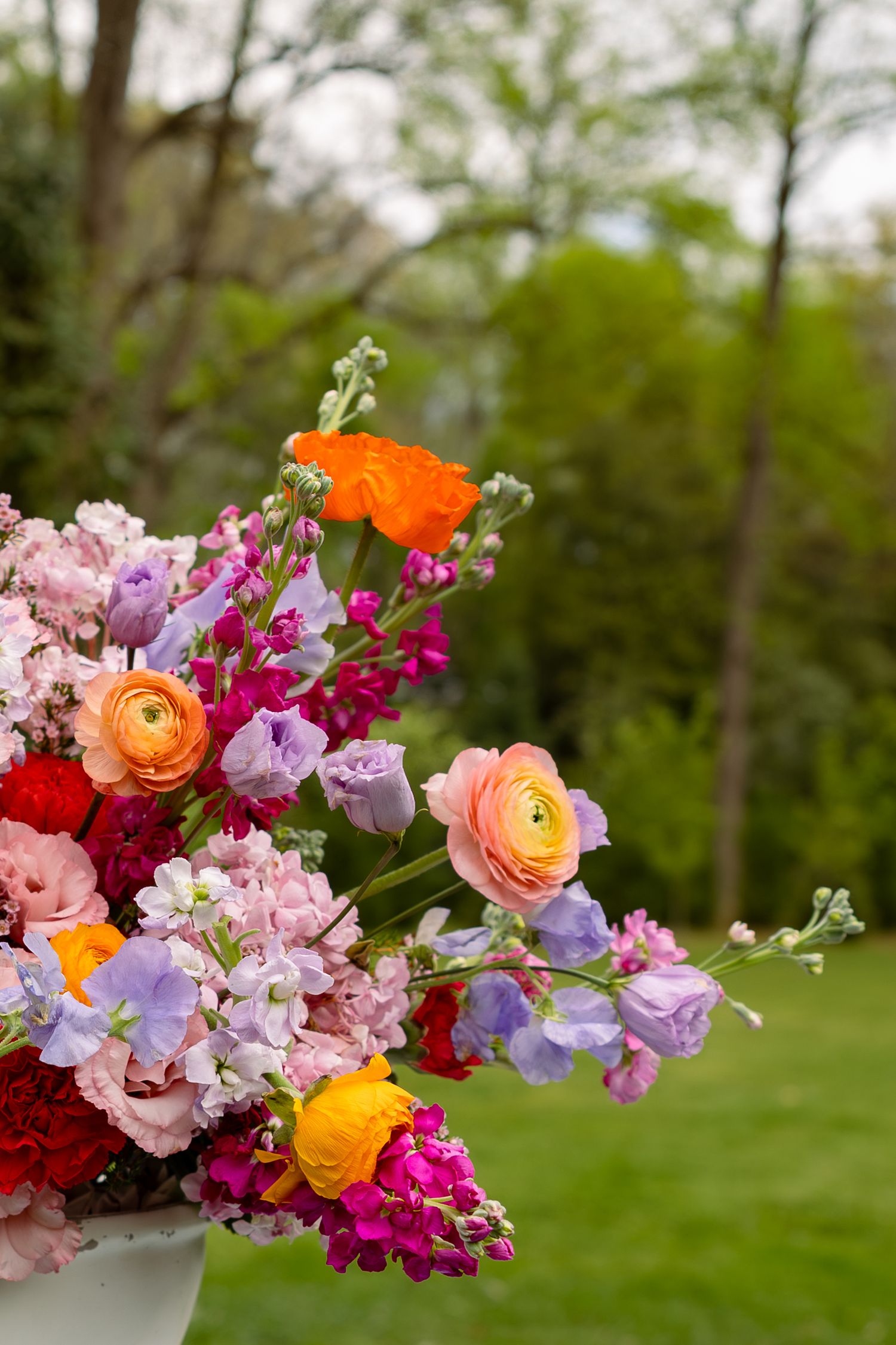 Vibrant spring bouquet with orange, pink, purple, and yellow ranunculus and sweet peas in an outdoor garden setting.