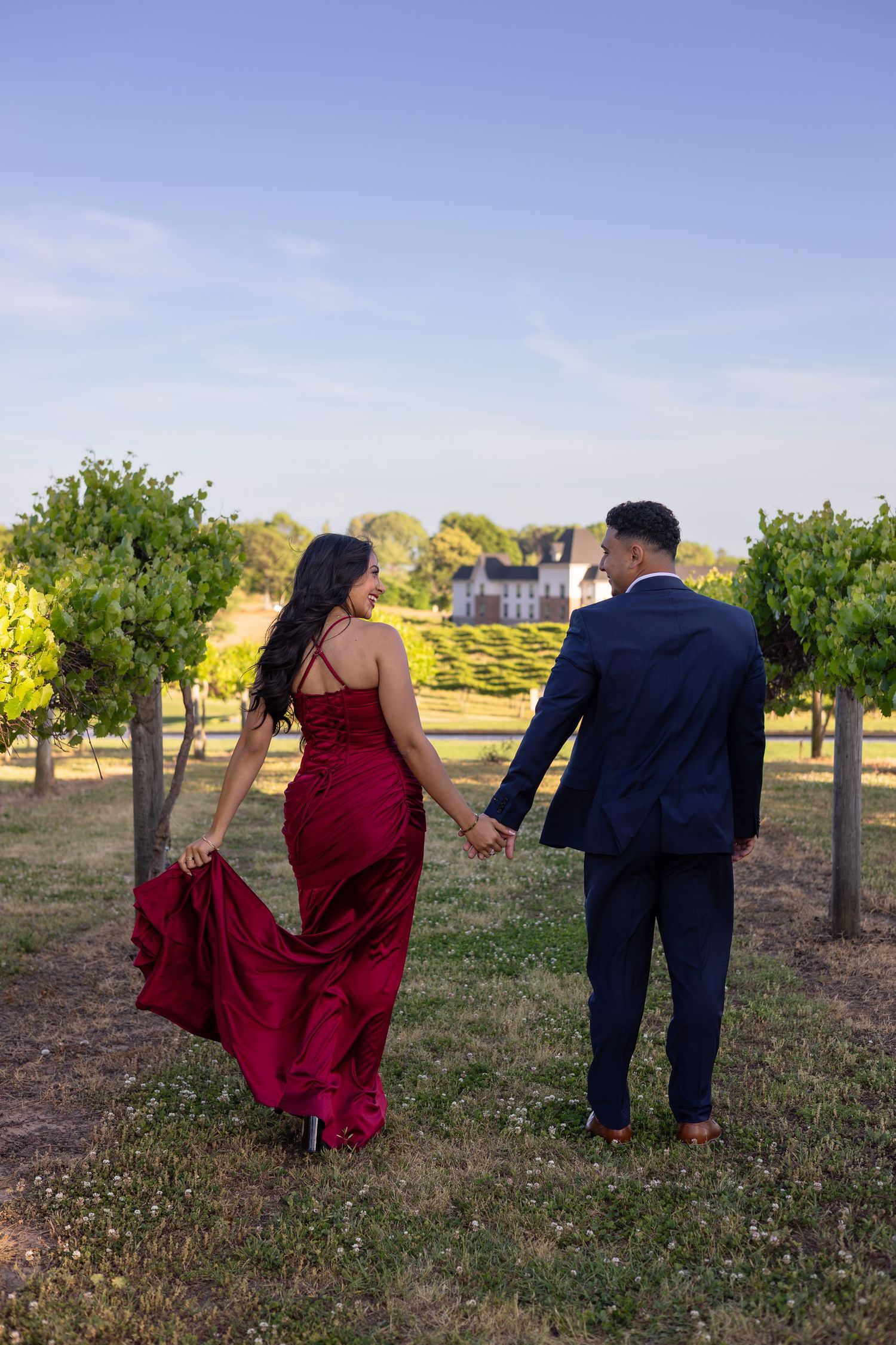 Couple in formal wear walking hand-in-hand through a vineyard, woman in red gown, man in navy suit, estate in background.