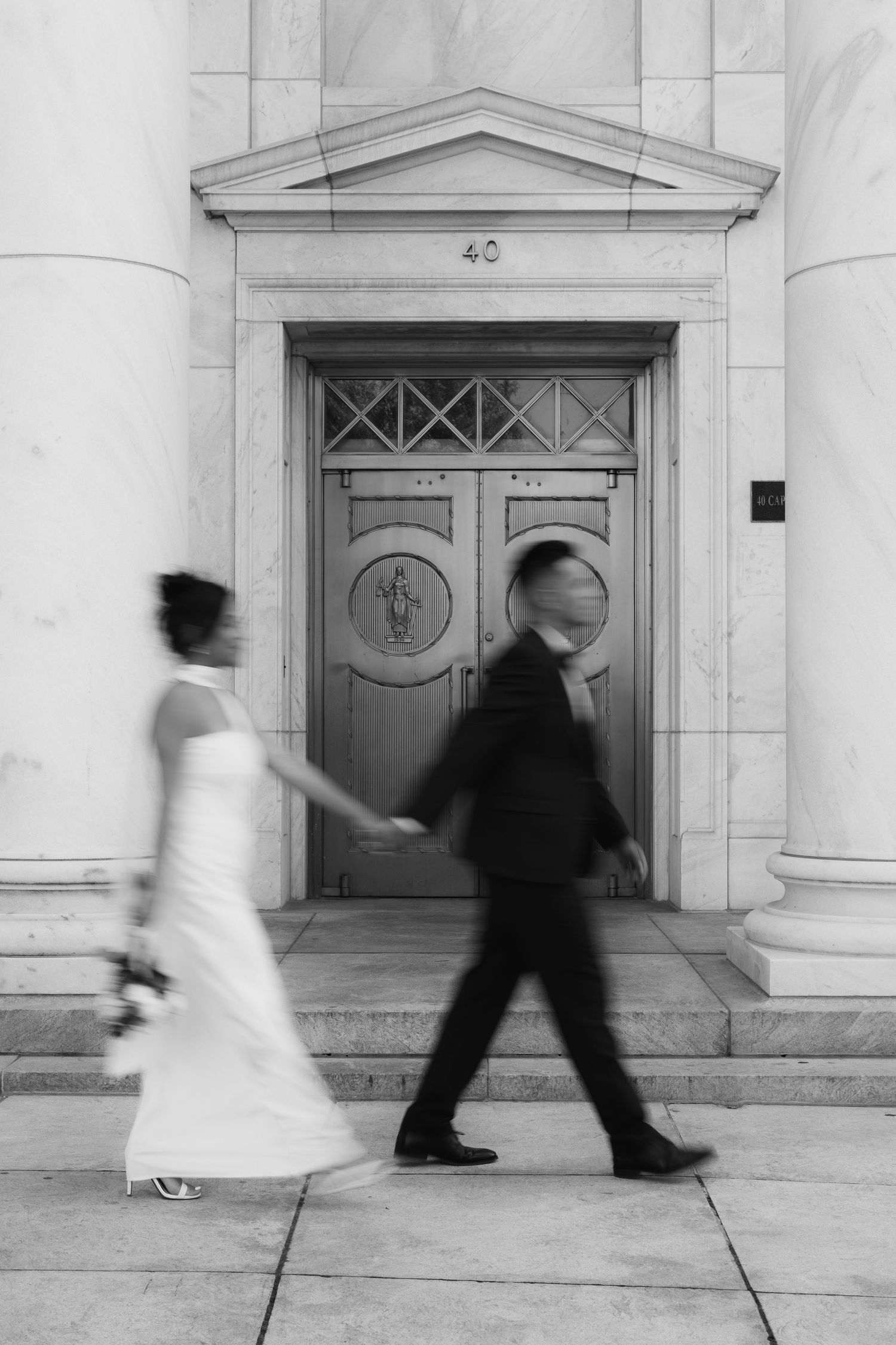 Black and white photo of a bride and groom walking hand in hand past an ornate door, motion blur capturing their joyful stride.