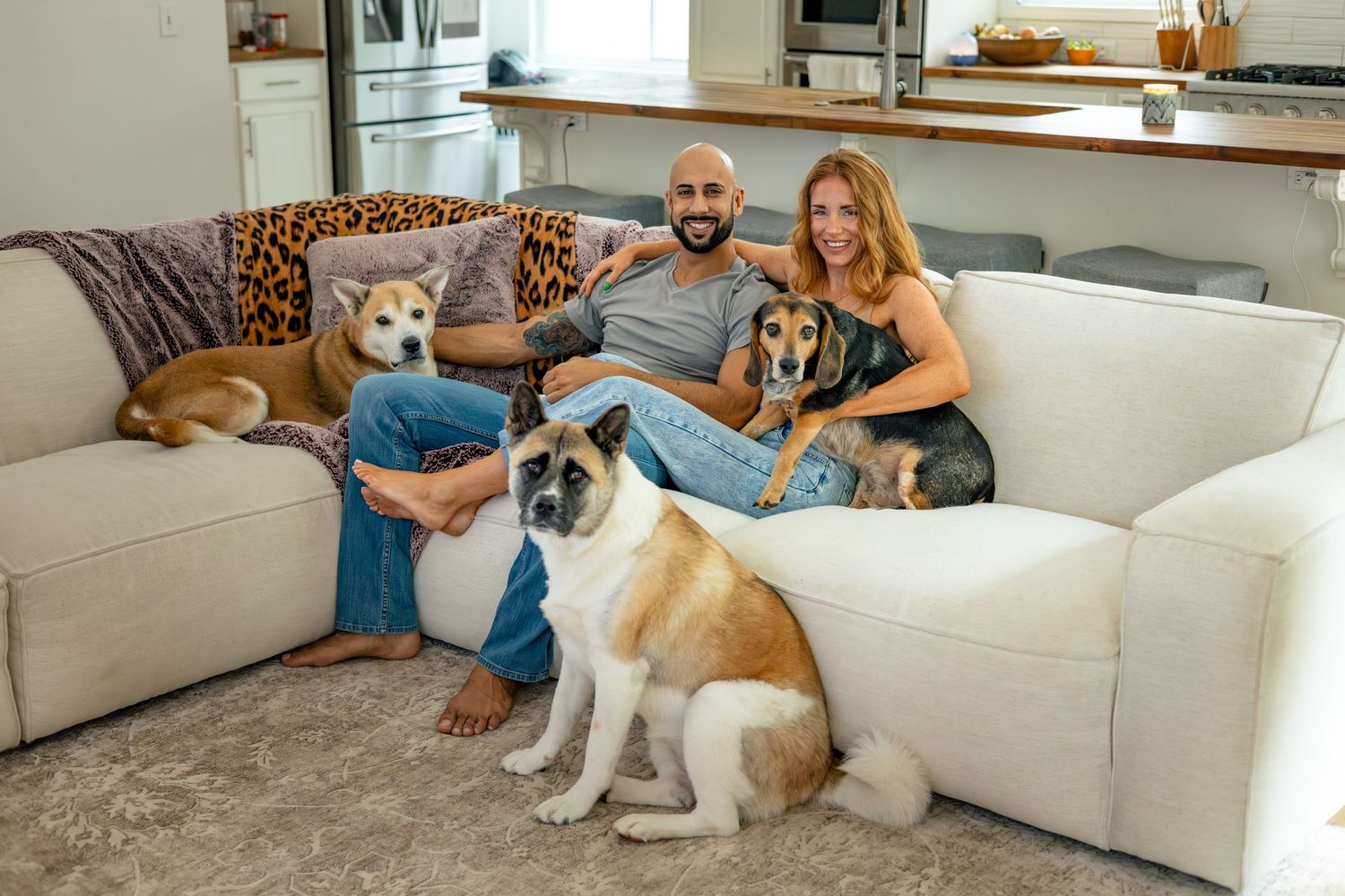A couple relaxes on a white couch with their collection of dogs.
