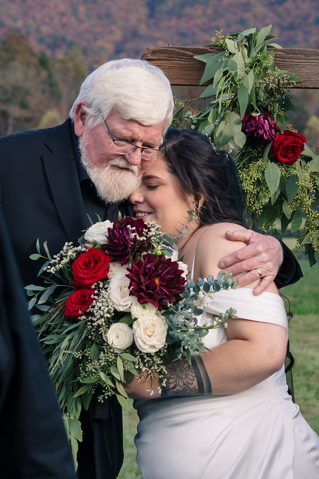 Wedding couple embraces with red and white floral bouquet against mountain landscape.