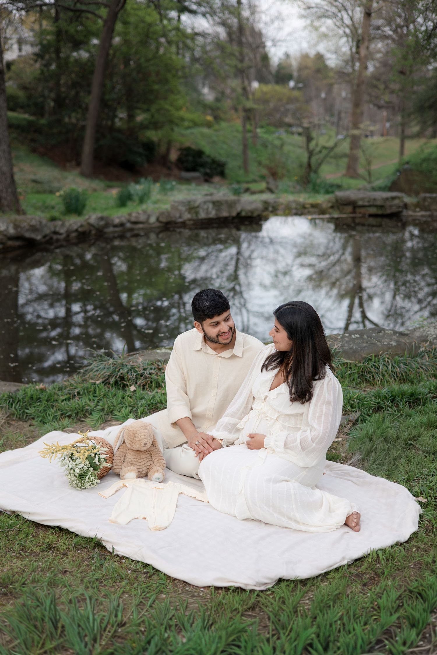 A couple in white outfits having a romantic picnic by a serene pond with spring trees in the background.