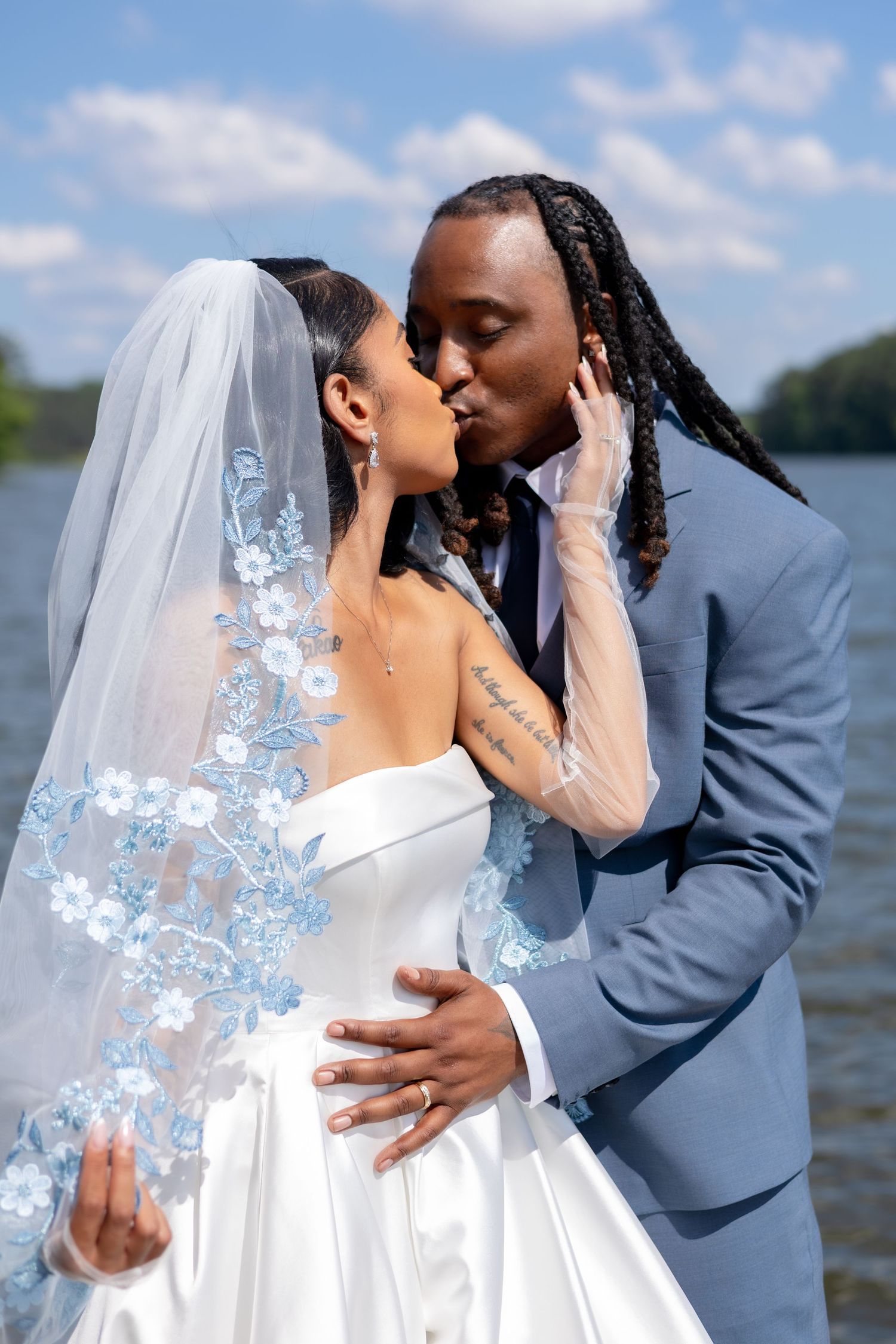 Interracial couple sharing a kiss on their wedding day by a lake, bride in white gown with blue floral veil.