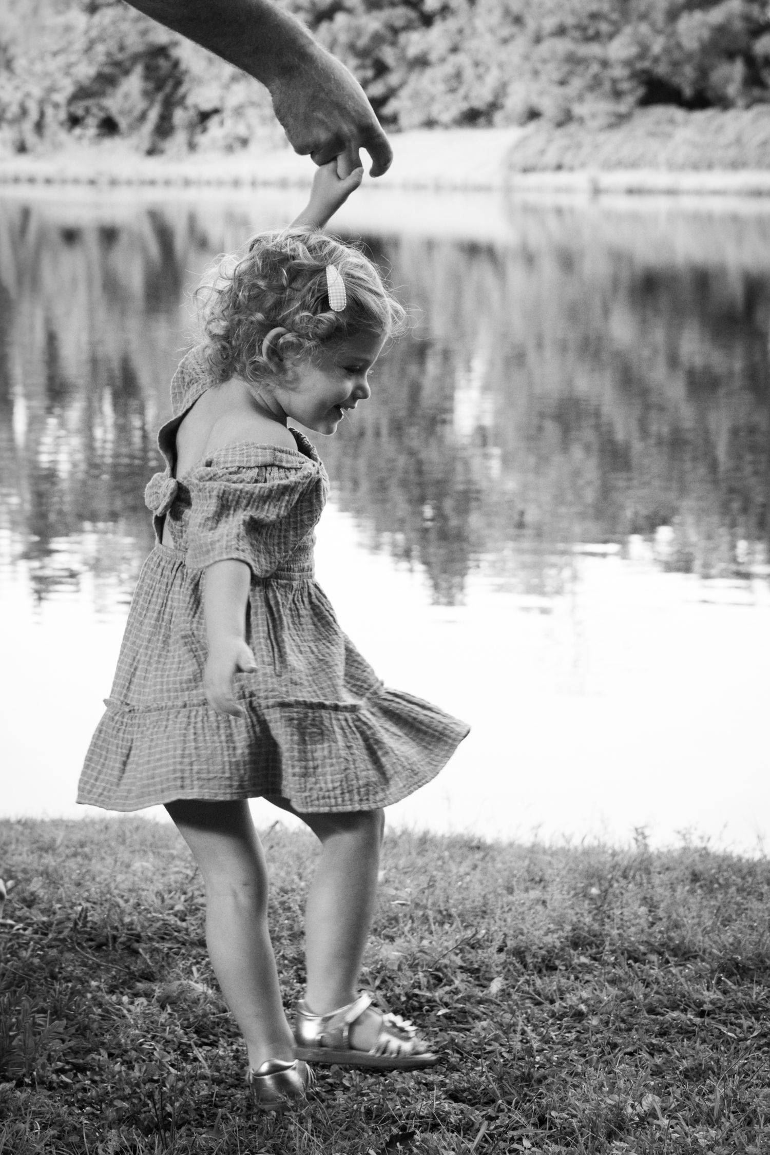 A young child in a gingham dress twirls by a lake in black and white.