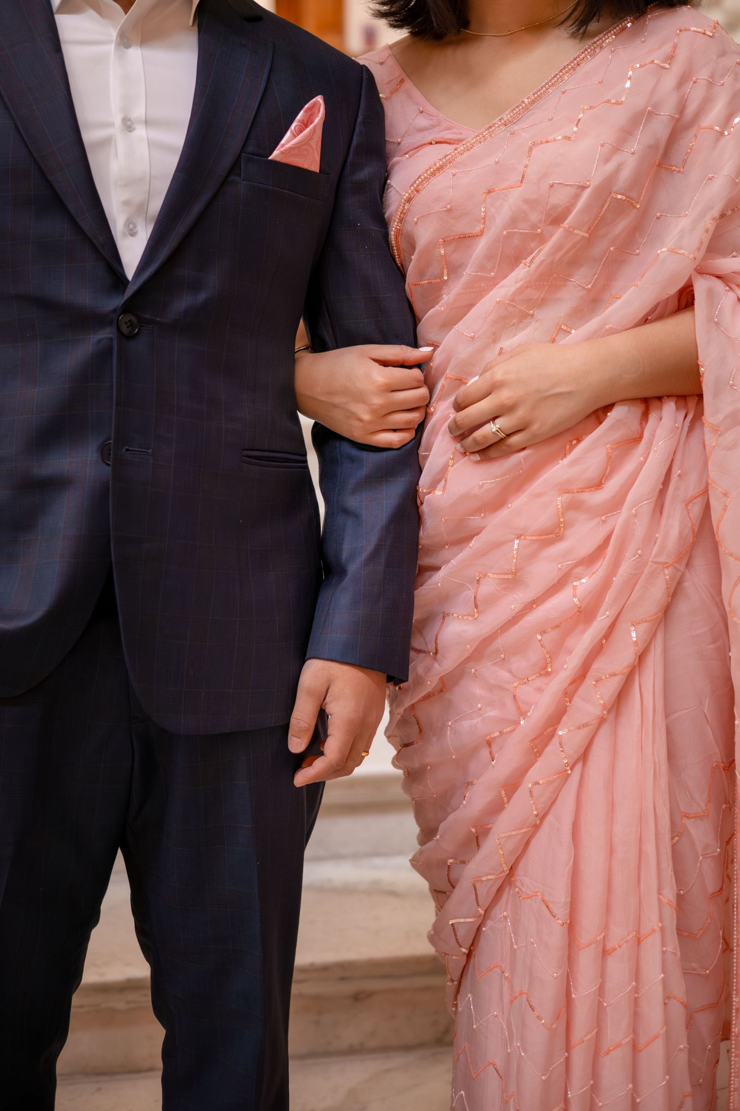A man in a dark suit stands next to a woman wearing a pink and white patterned saree.