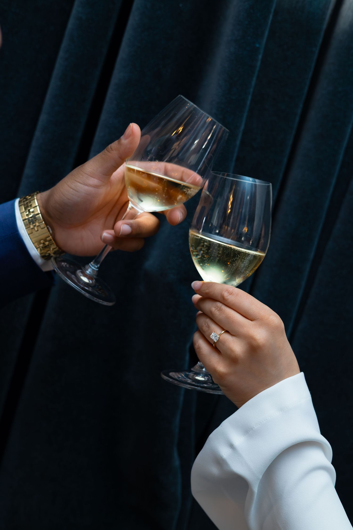 Two hands clinking wine glasses filled with white wine, one wearing a gold watch and diamond engagement ring.
