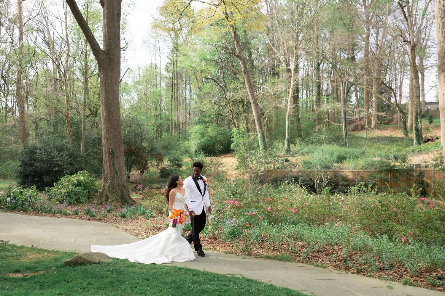 Bride and groom walk hand-in-hand on a garden path surrounded by lush greenery and trees on their wedding day.
