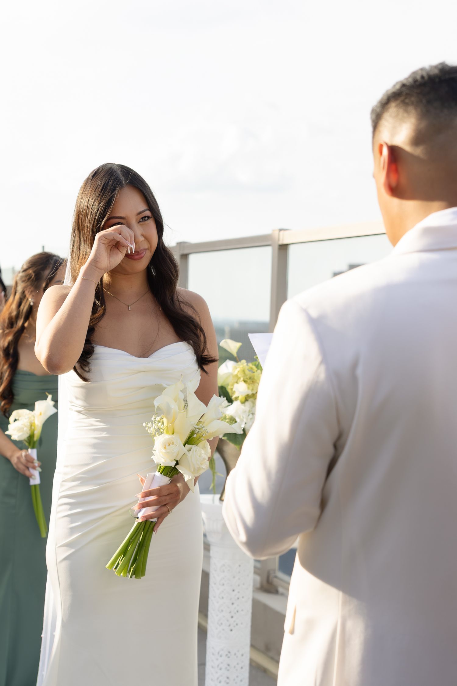 Emotional bride wiping tears holding white bouquet facing groom during outdoor rooftop ceremony.