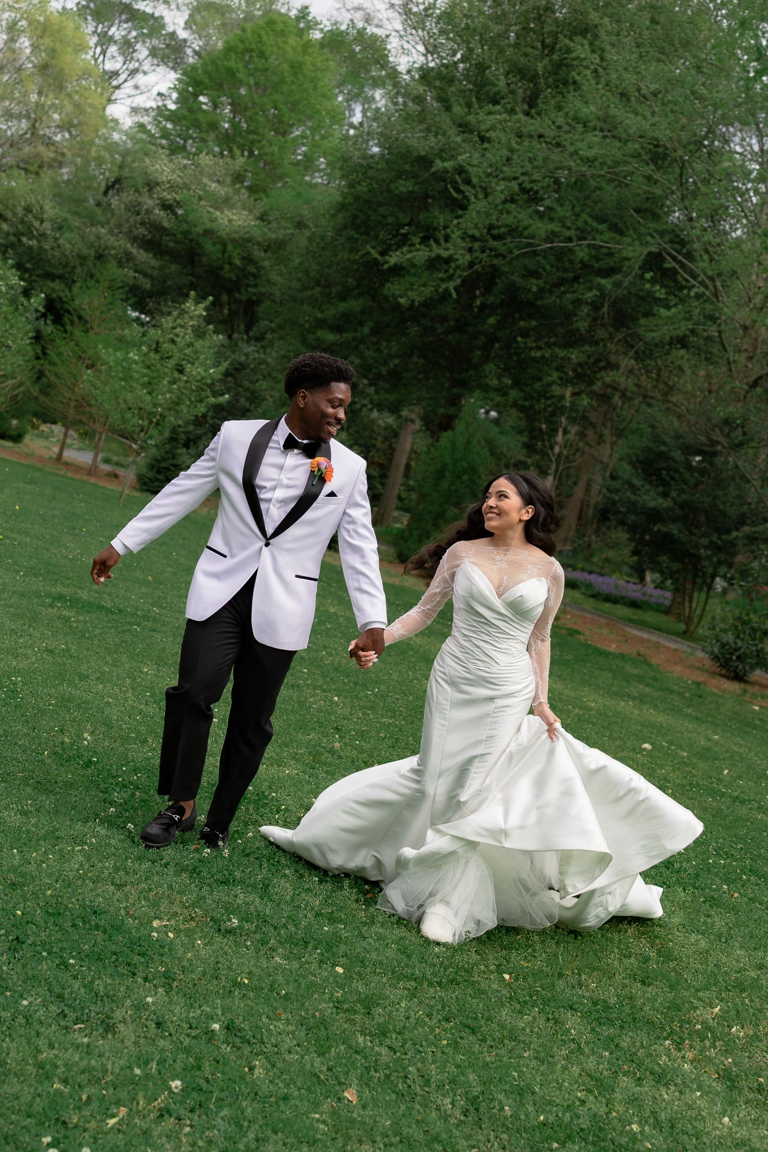A joyful bride and groom hold hands while walking across a lush green lawn in their elegant wedding attire.