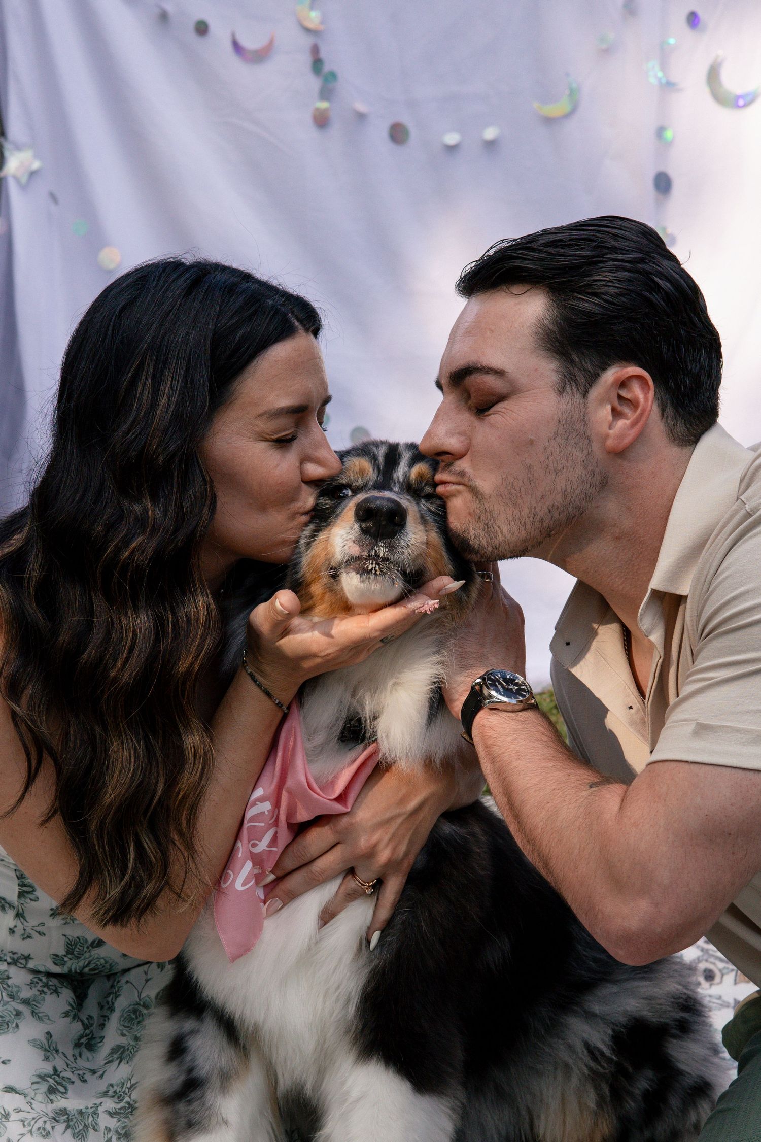 A couple kisses their dog from both sides while sitting on a decorated backdrop.