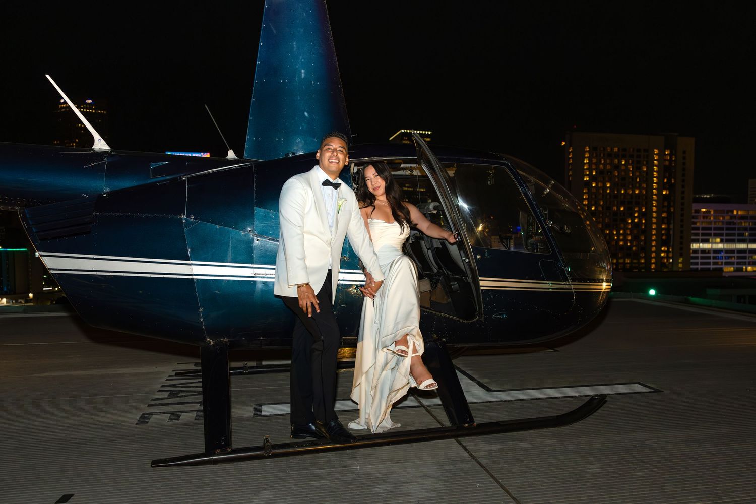Bride and groom posing in front of a helicopter at night with city lights in the background.