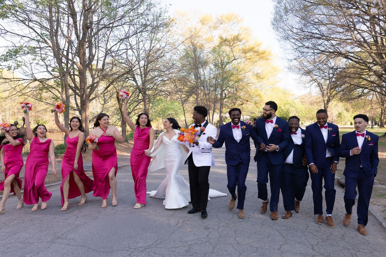 Wedding party walks outdoors, bridesmaids in pink gowns and groomsmen in navy suits, celebrating with the bride and groom.