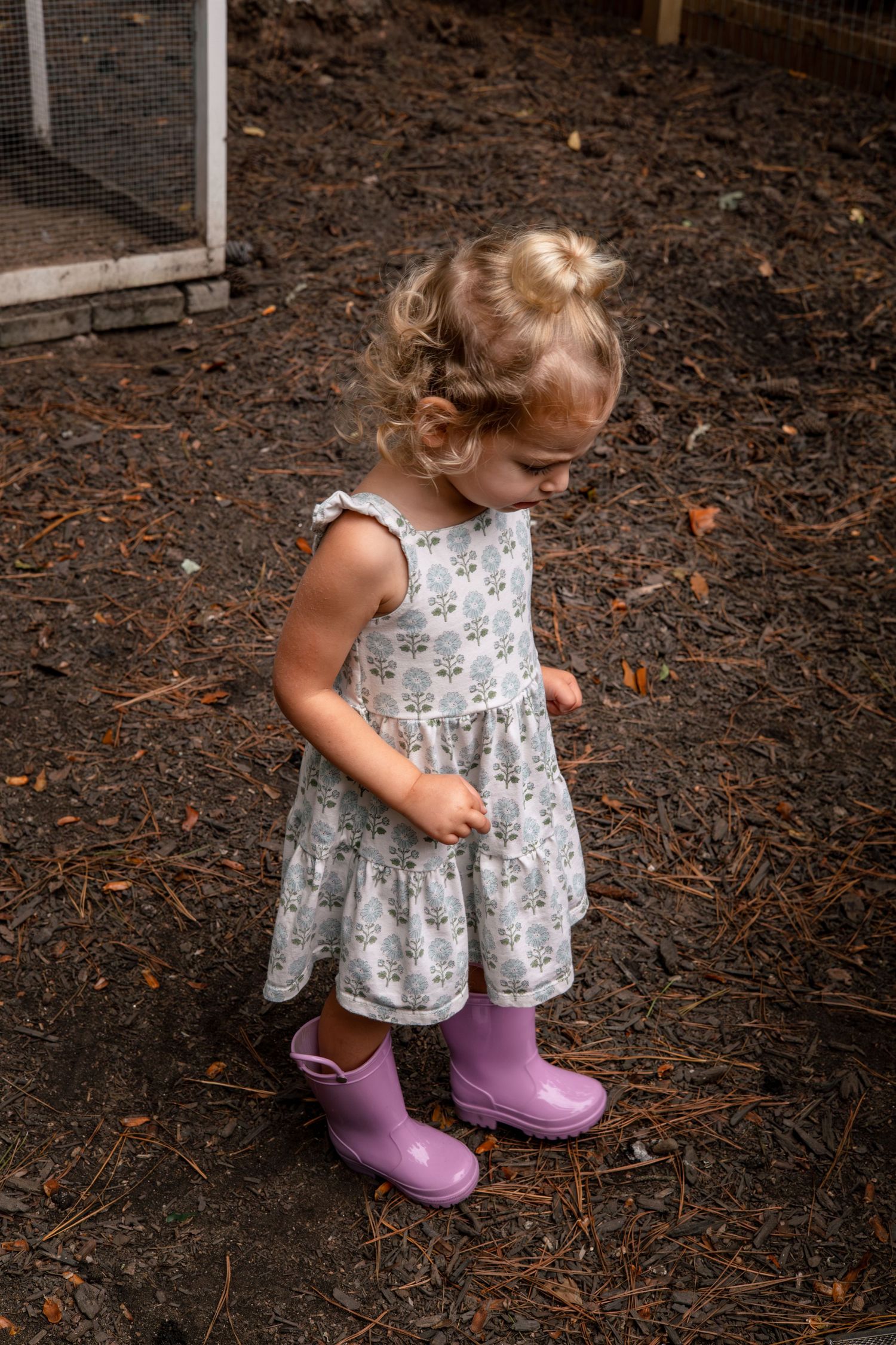A small child wearing a floral dress and purple rain boots explores mulch-covered ground.
