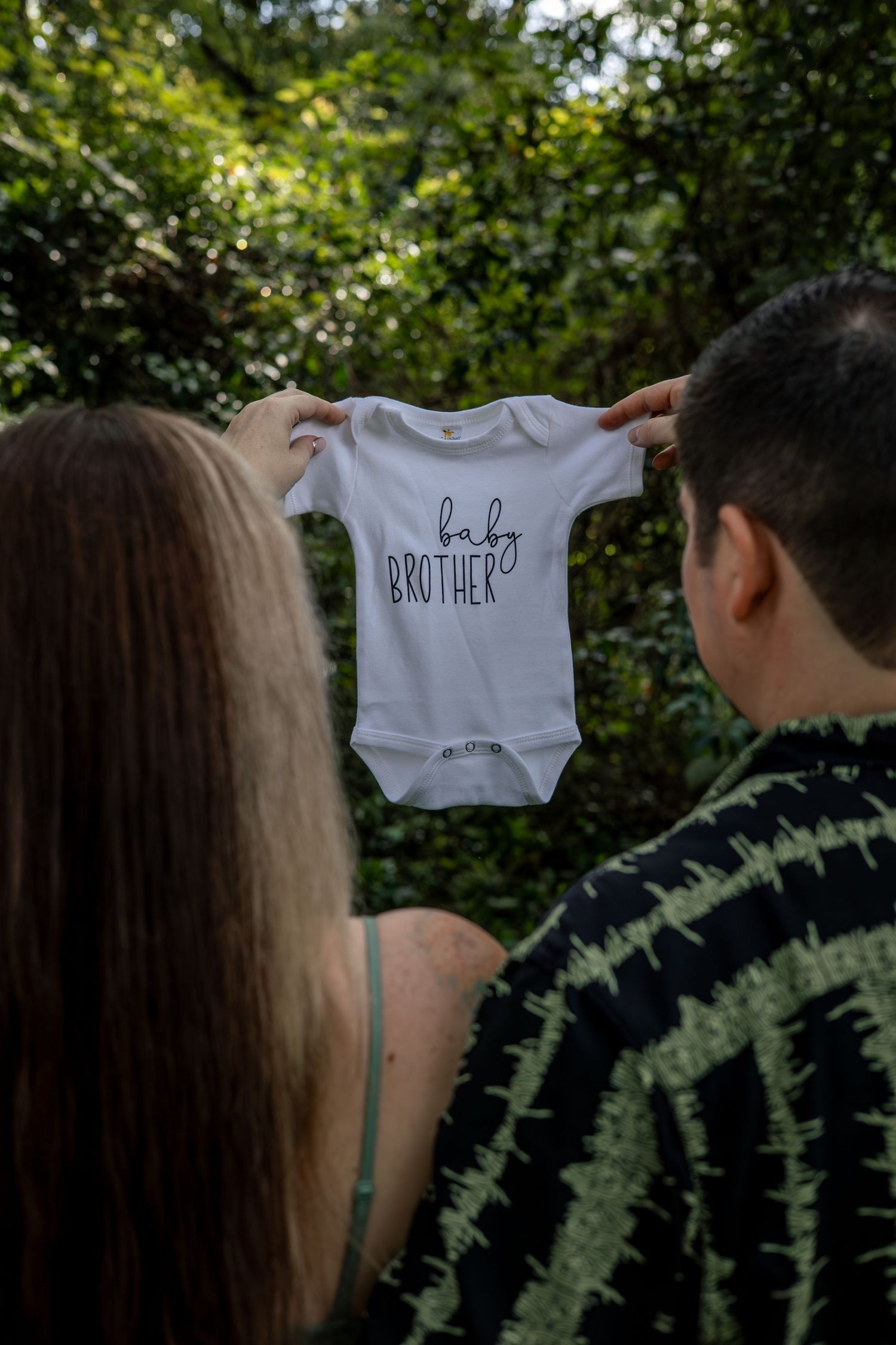 A couple holds up a white baby onesie with 'baby brother' text in an outdoor setting.
