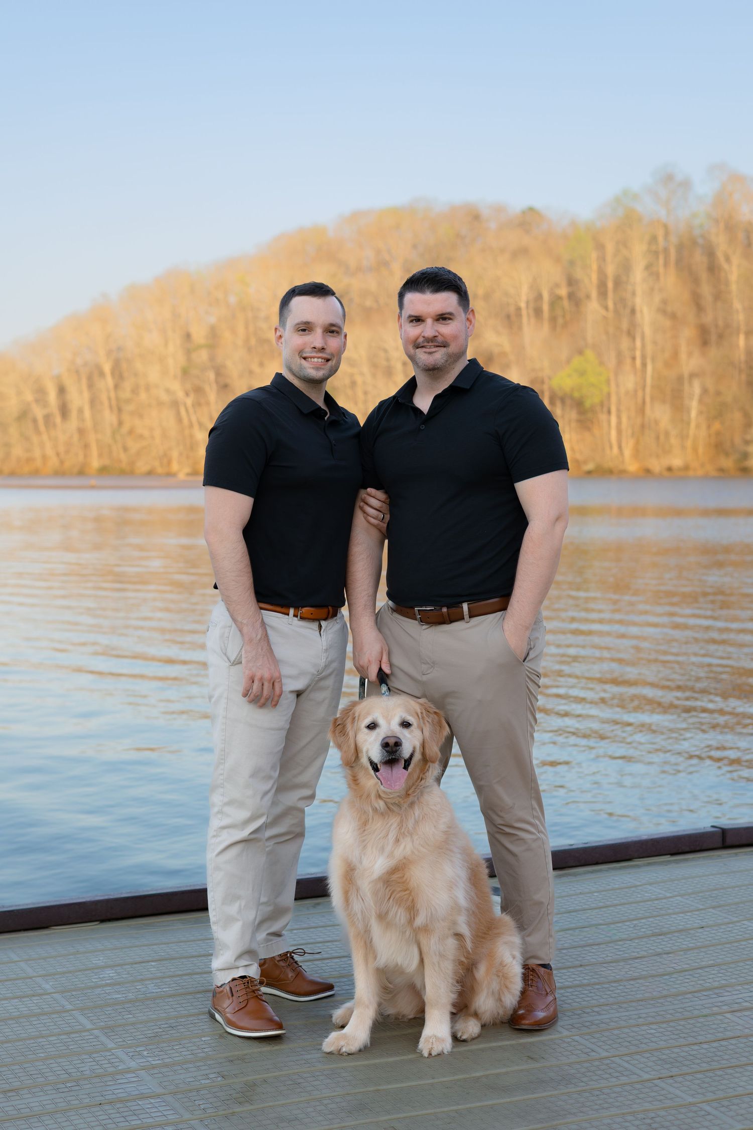 A couple poses with their golden retriever on a wooden dock by a serene lake during golden hour.