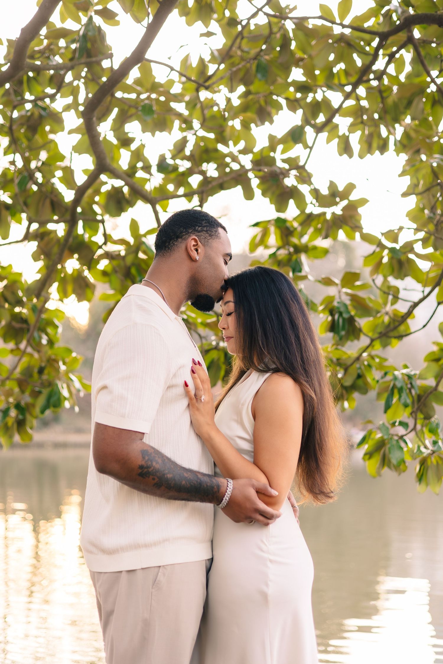 Romantic couple in white outfits sharing tender moments by a lake with tree branches overhead during golden hour.