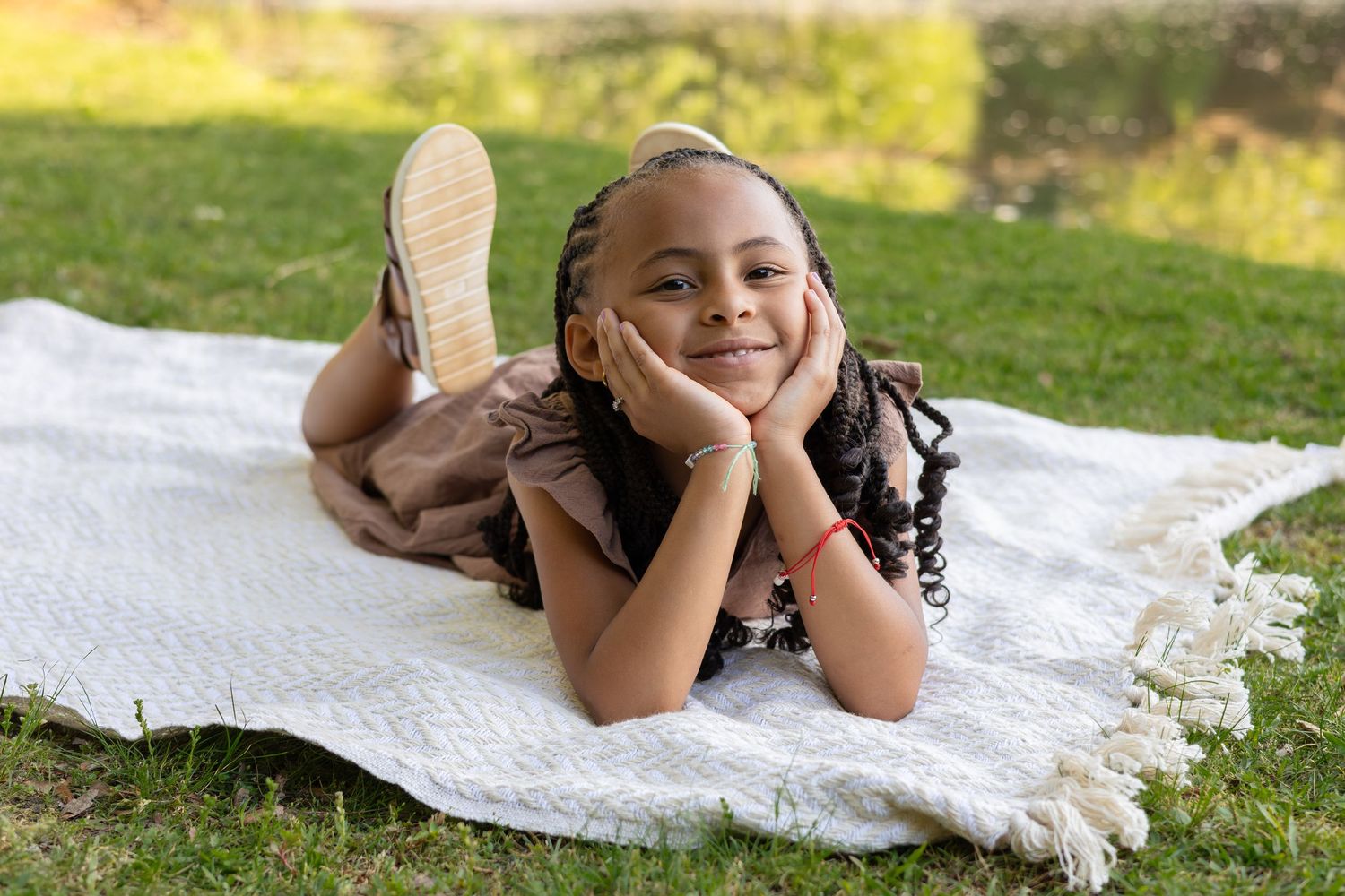 A young girl with braids lies on a white blanket on grass, smiling with hands on cheeks.
