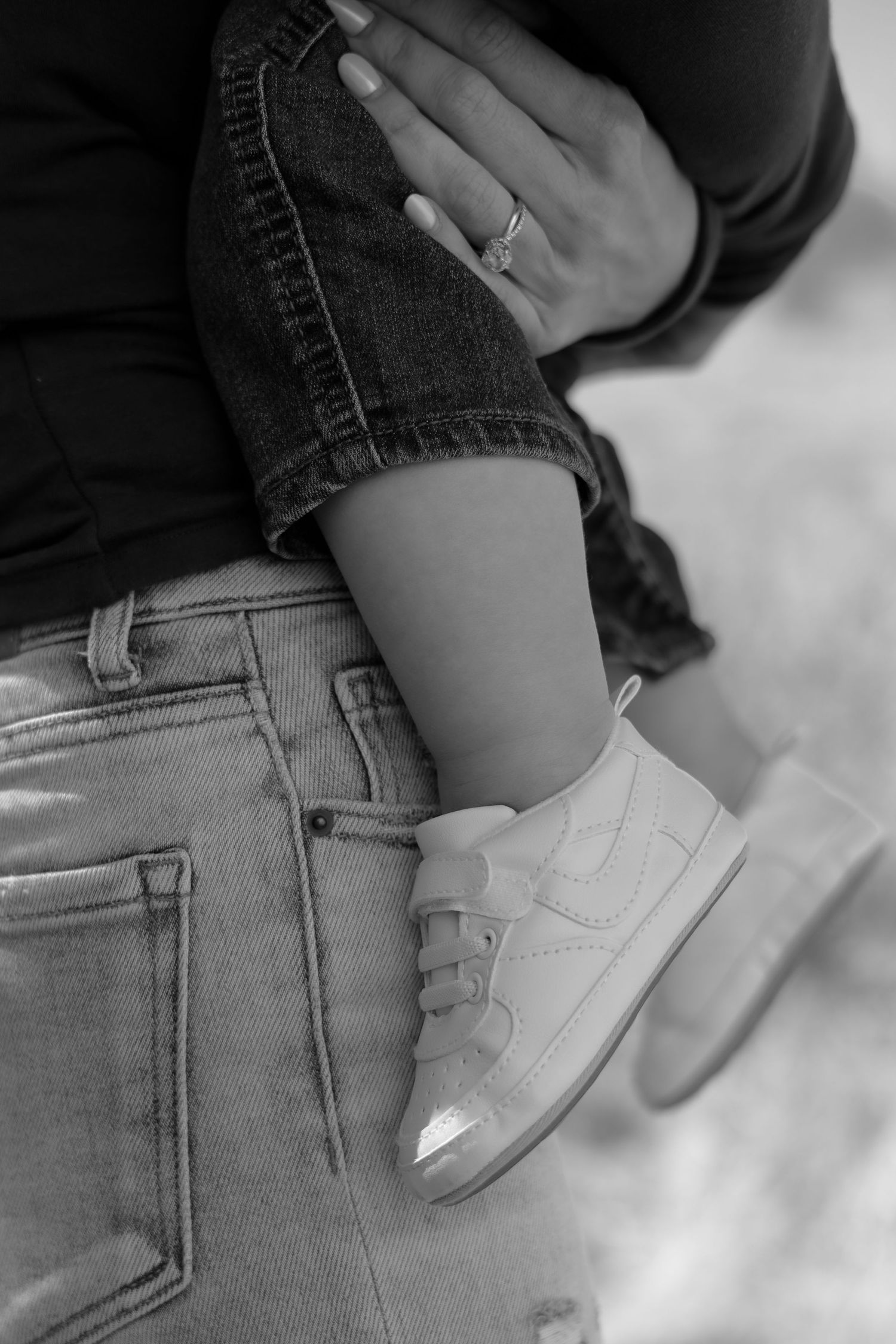 Black and white photo of a mother holding her baby wearing white sneakers, with a ring visible on her hand.