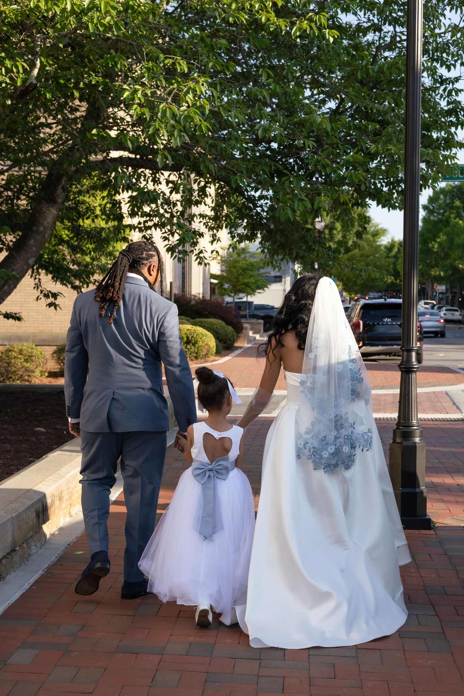 Bride and groom in wedding attire walk hand-in-hand with flower girl in white dress on brick sidewalk.