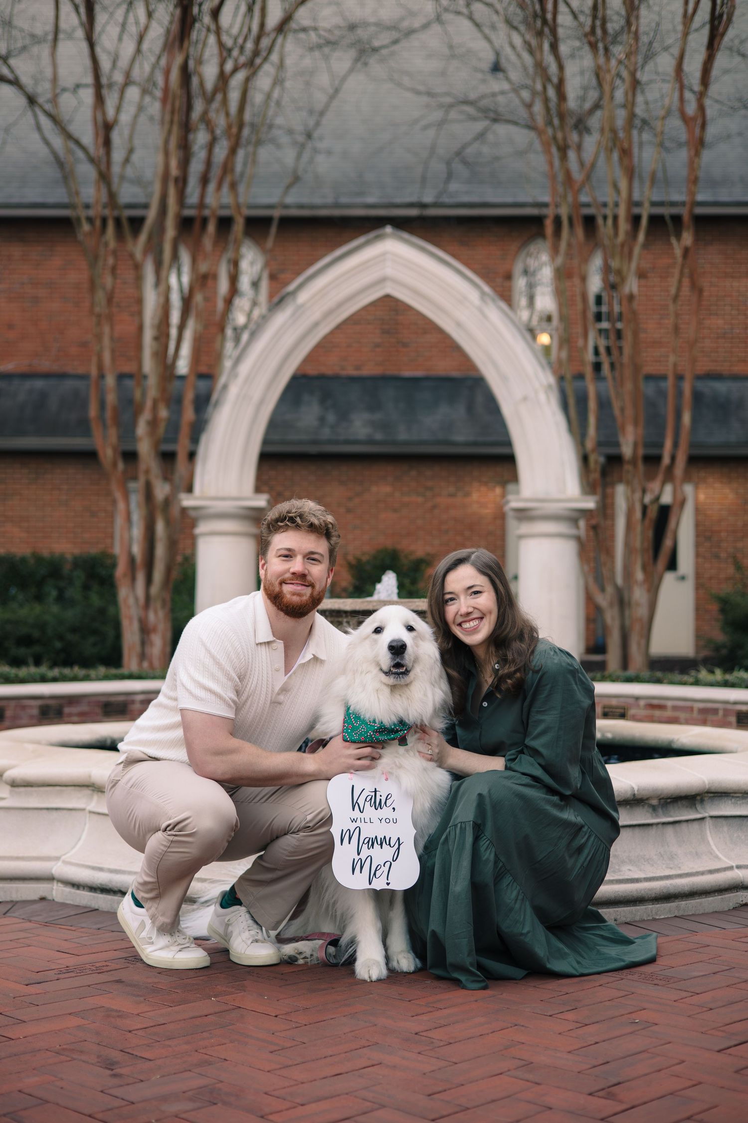 A couple sits with their white dog by a fountain holding engagement announcement signs.