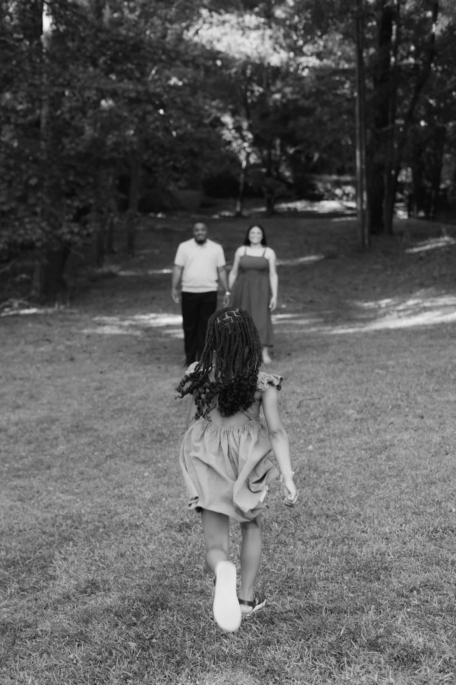 Black and white photo of a girl running toward her parents in a lush green park setting.