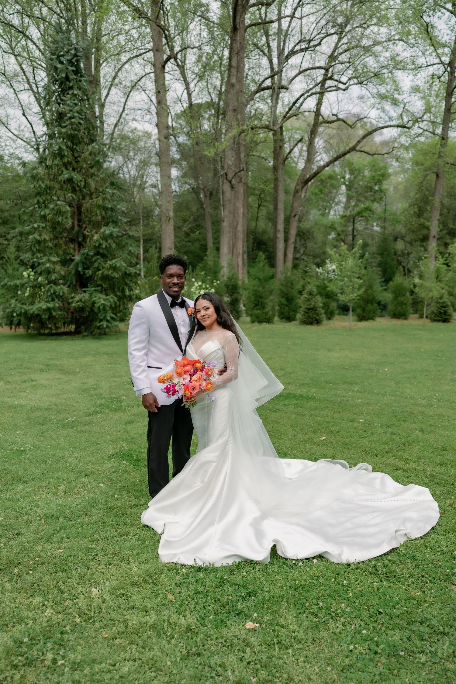 Bride and groom pose on green lawn, she wears a white gown with train, he wears a white tuxedo jacket.