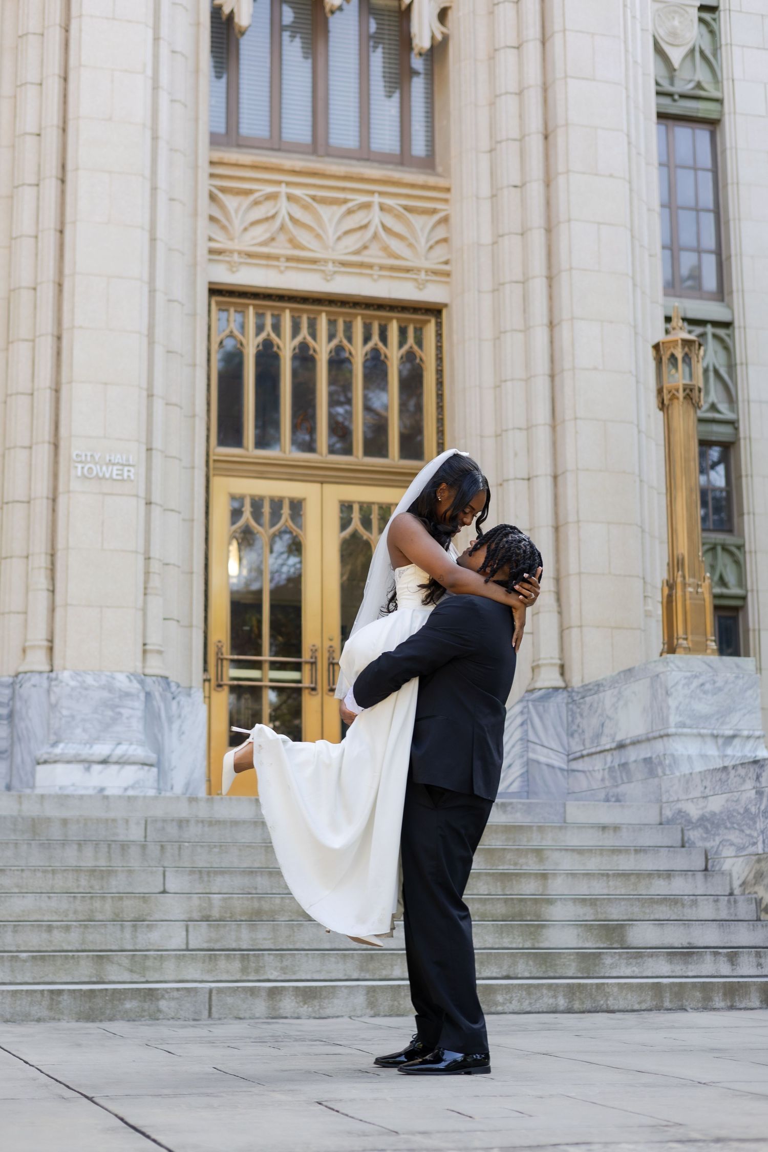 Groom lifting bride in white gown outside ornate church steps on their wedding day.