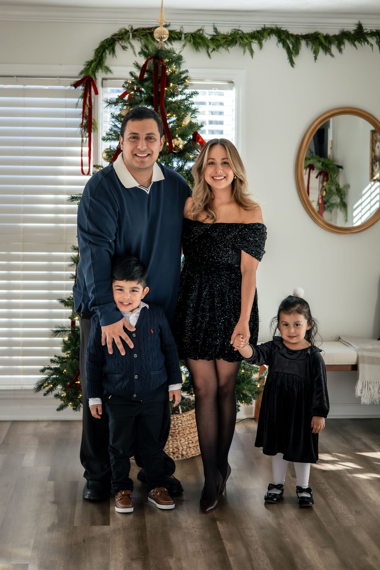 Family dressed in black poses together by Christmas tree in living room with holiday decorations.