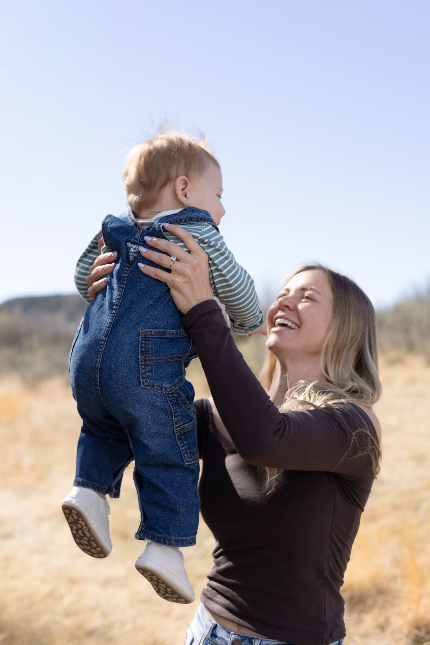 Happy mother lifting smiling baby in denim overalls outdoors on a sunny day in a dry field.