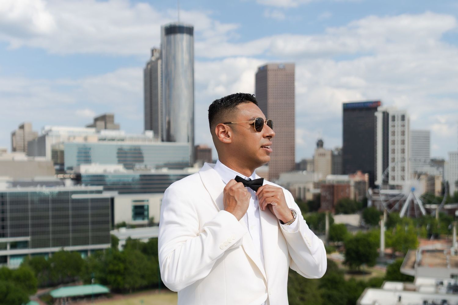 Man in white tuxedo adjusting bow tie with city skyline backdrop on a cloudy day.
