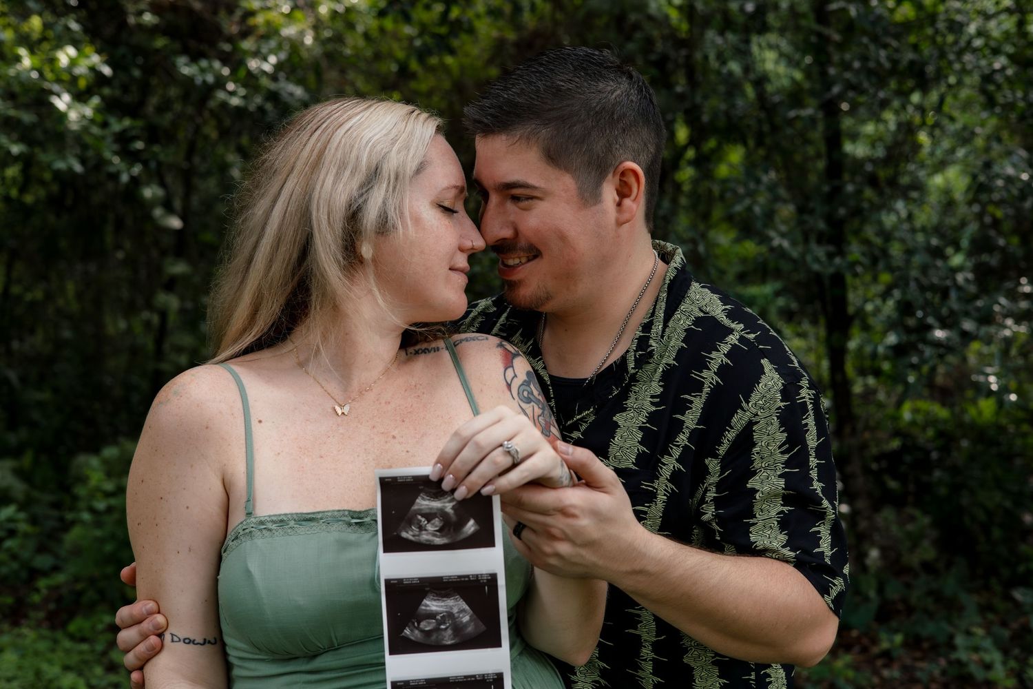 A couple shares an intimate moment while holding ultrasound photos outdoors.