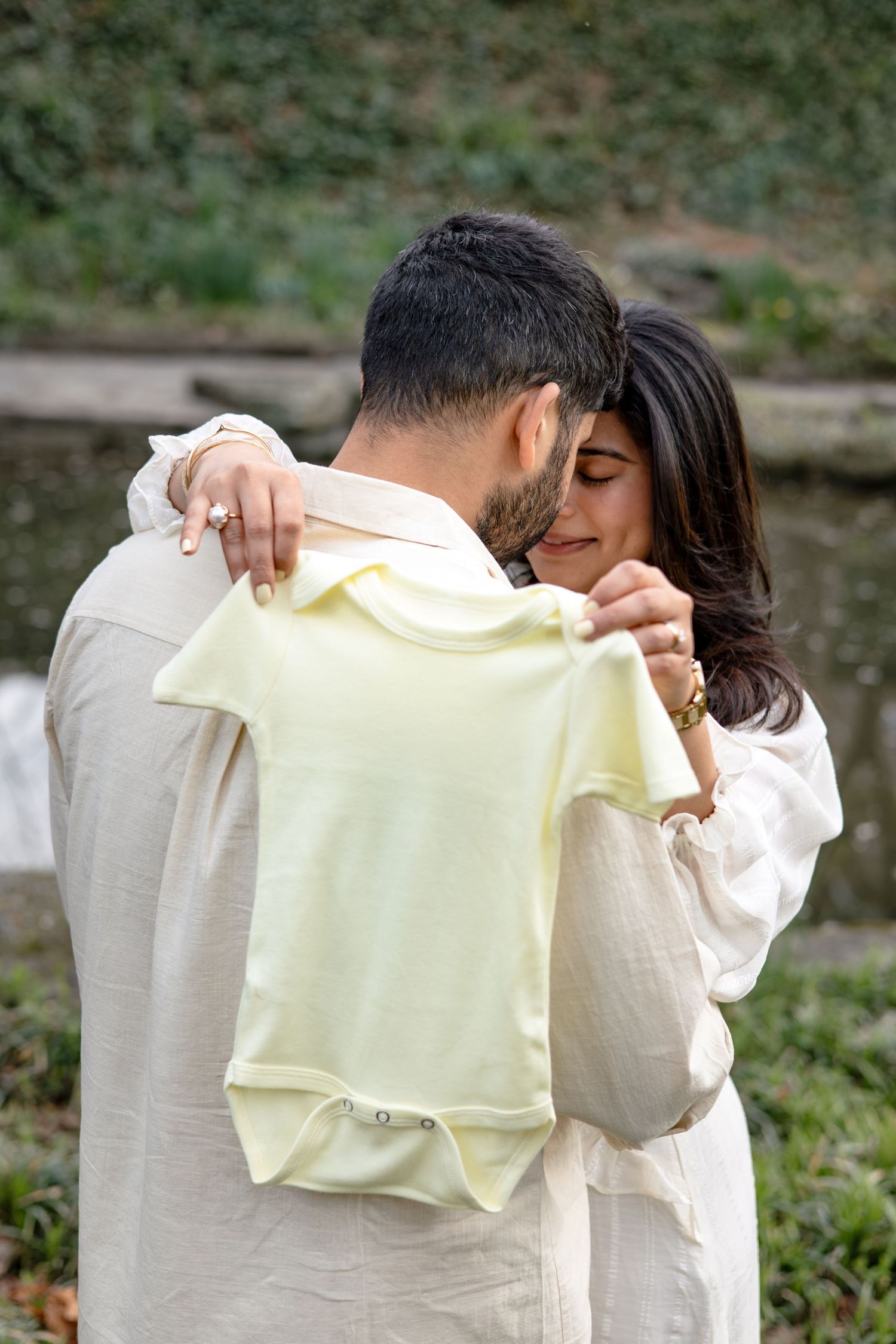 A couple embraces while holding a small yellow baby onesie outdoors in a tender moment.